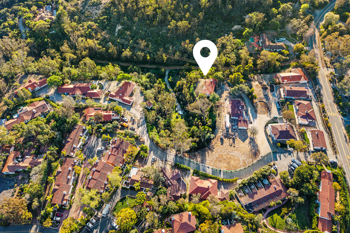 An aerial view of a residential neighborhood with houses, trees, and a school yard with a basketball court. A location pin marks a house surrounded by greenery.