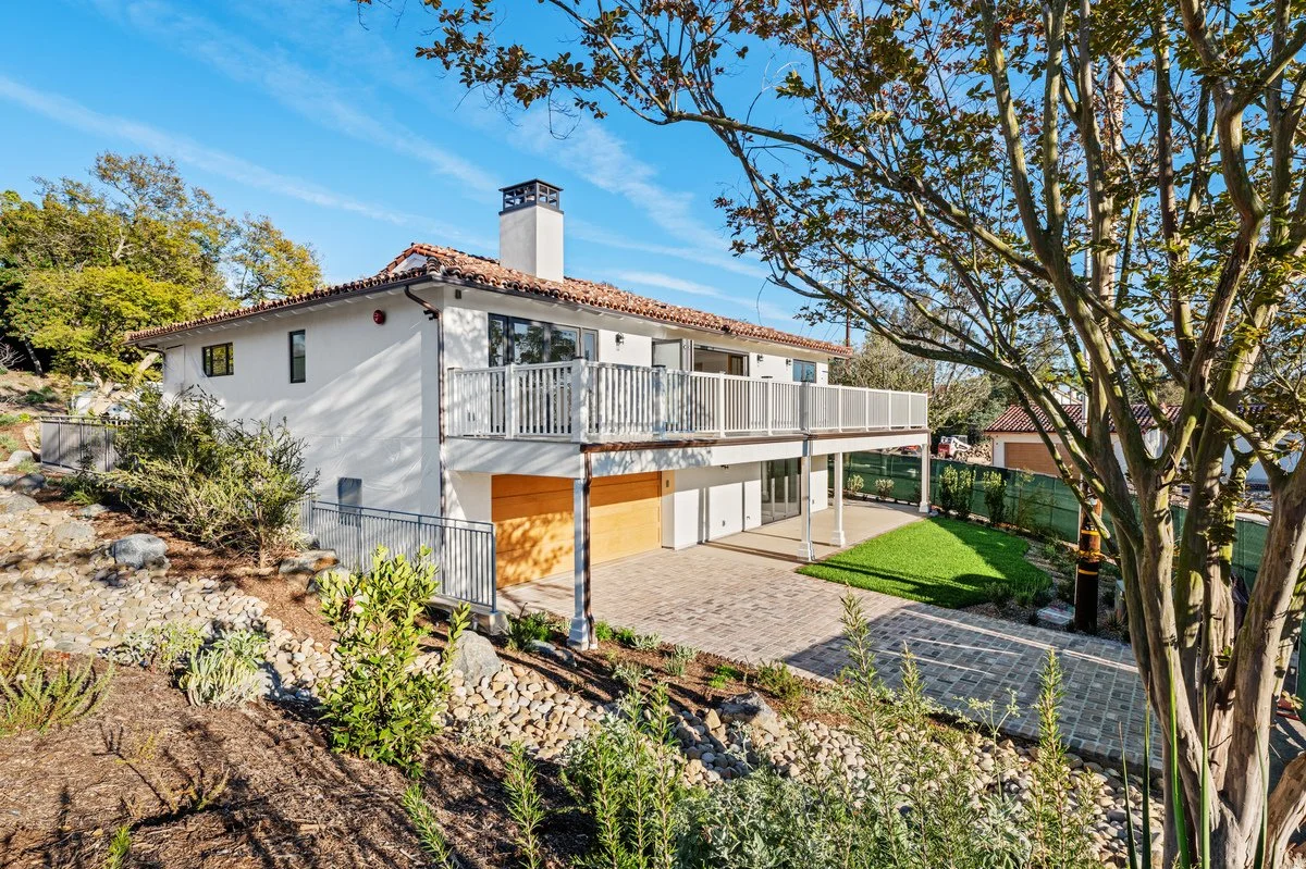 Front view of a modern two-story house with a white exterior, red tile roof, and a large balcony on the upper floor. The house has a paved driveway, a small lawn, and landscaped garden with rocks and plants, surrounded by trees and a green fence under a blue sky.