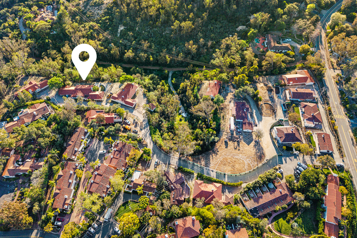 An aerial view of a residential neighborhood with houses featuring red-tiled roofs, surrounded by lush green trees. There is a small school or park with a basketball court in the center, enclosed by a fence. A winding road runs along the right side of the image, and a utility line cuts through the upper part of the scene. A white location marker is positioned over one of the houses on the left side.