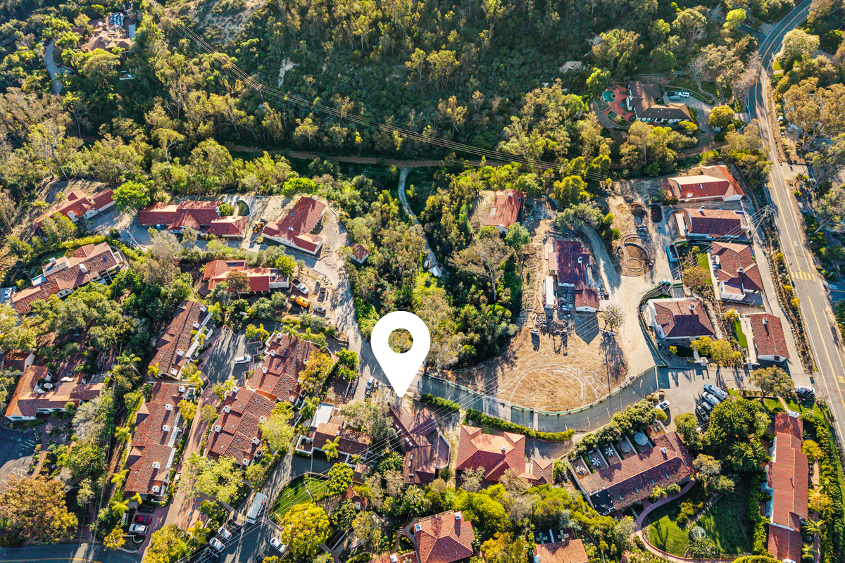 Aerial view of a neighborhood with houses, trees, and a schoolyard or playground area, with a white location marker indicating a specific spot.