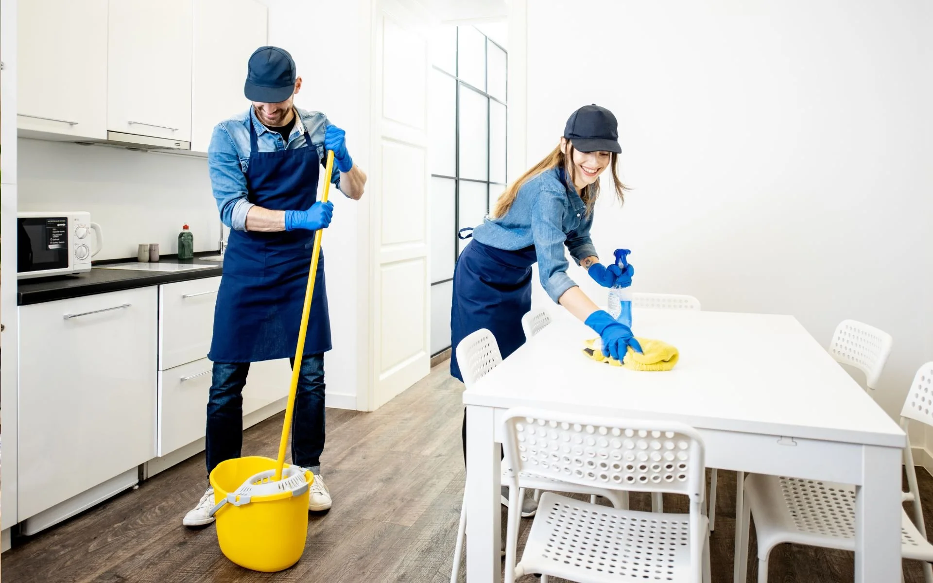 Two people cleaning a white dining table in a bright, modern kitchen, both wearing blue aprons, gloves, and caps. One person is mopping the floor, and the other is wiping the table with a yellow cloth.