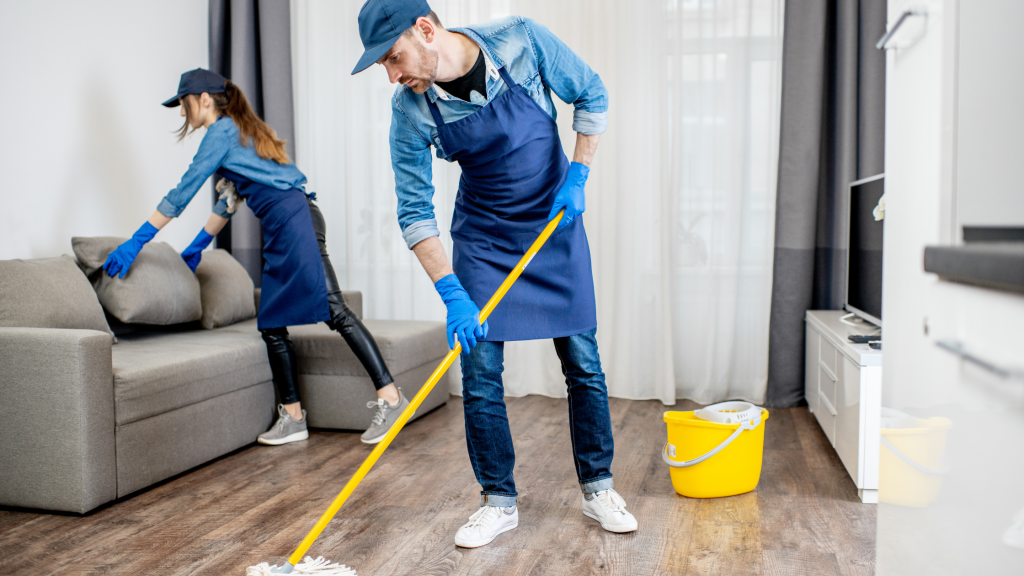 Two people cleaning a living room with yellow cleaning buckets, wearing blue aprons, gloves, and hats; a man mops the floor while a woman arranges pillows on the sofa.