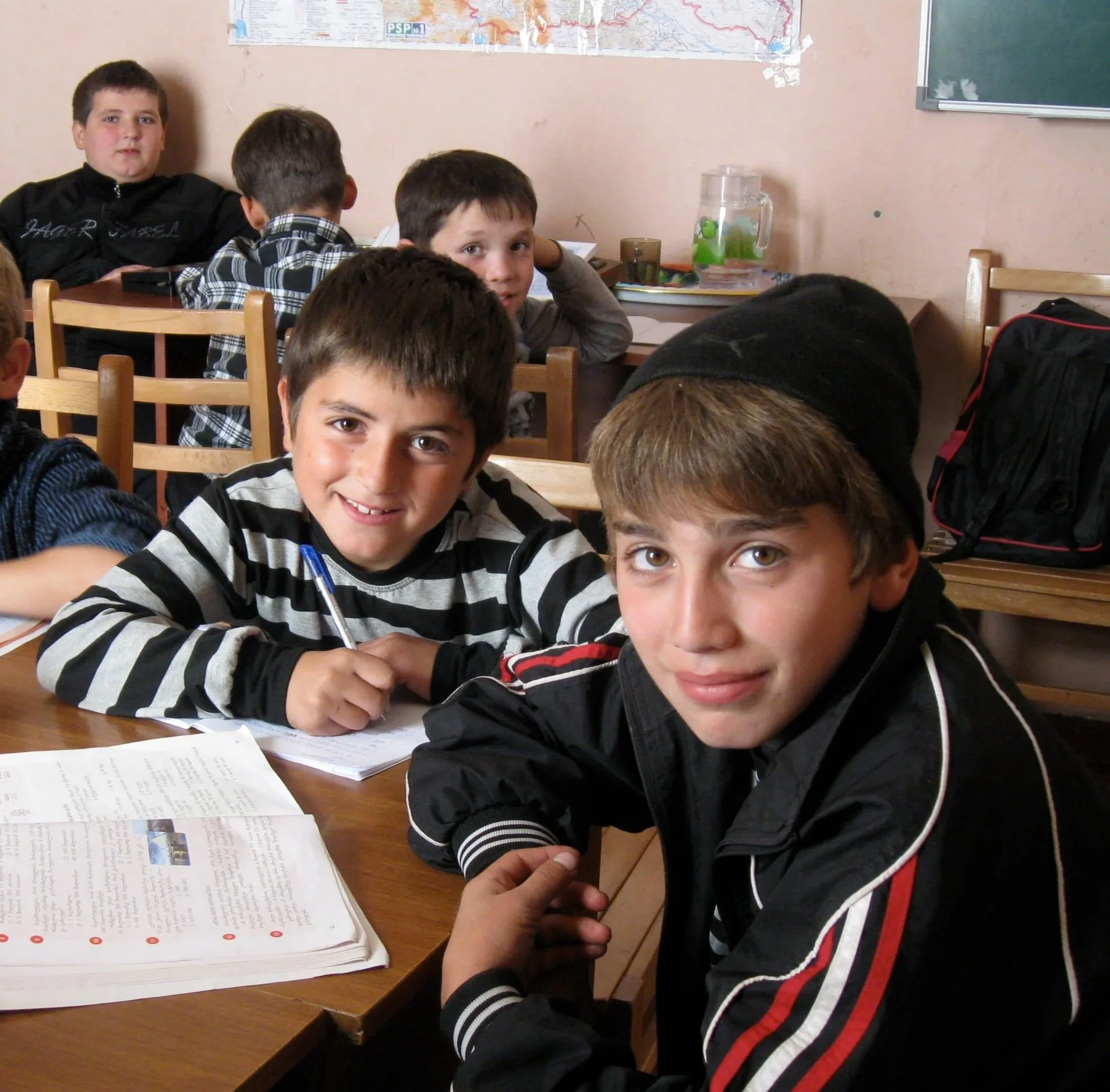 A group of young boys sitting at a table in a classroom, smiling at the camera. There are notebooks and papers on the table. The classroom has a map on the wall and a water pitcher in the background.