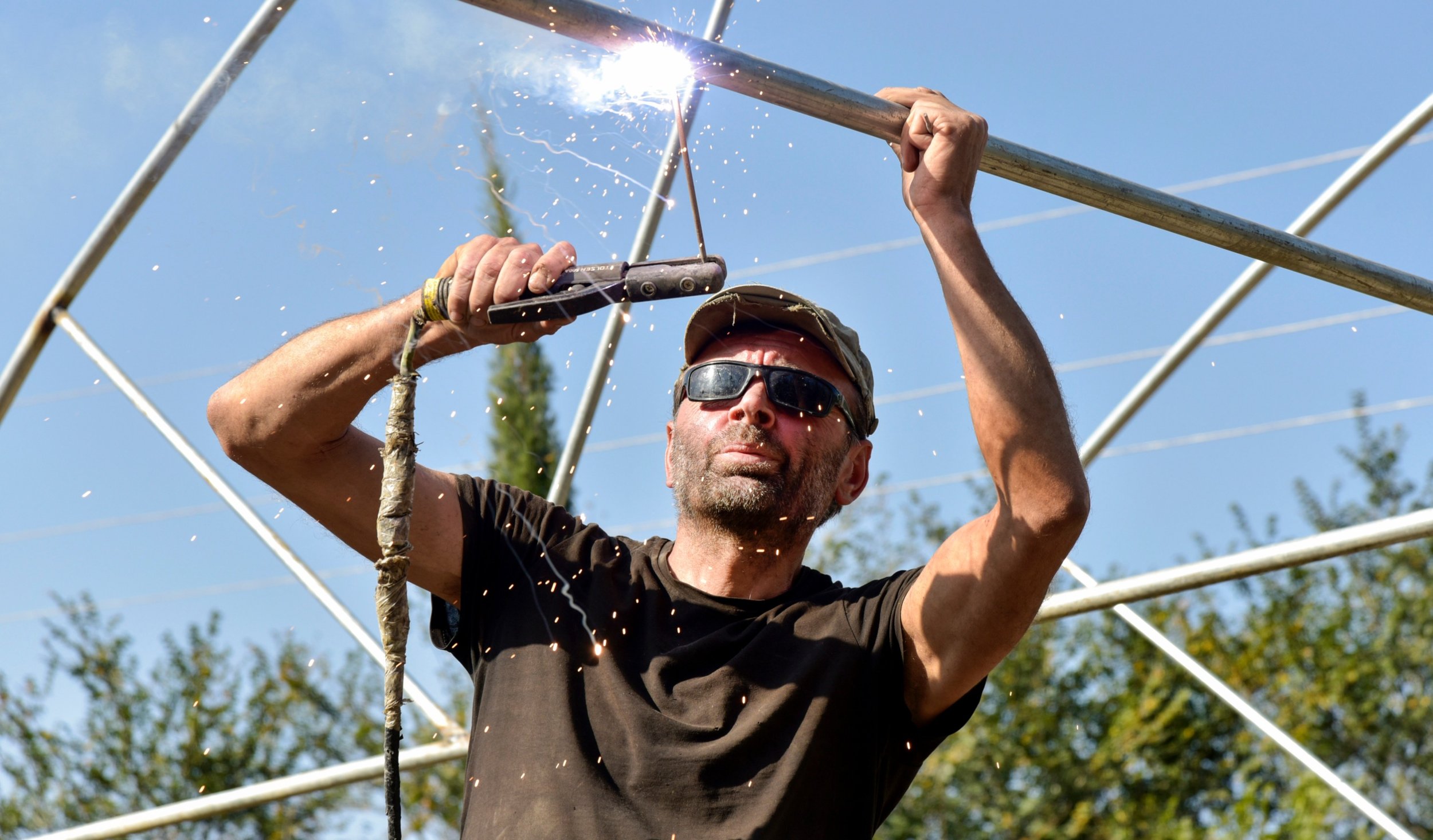Man wearing sunglasses and a cap using a metal tool to cut or grind a metal pipe outdoors during the daytime.