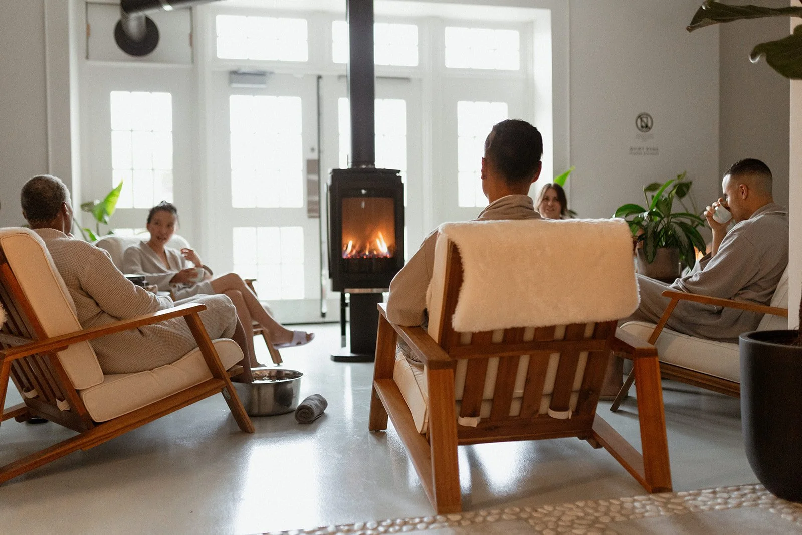 People relaxing in a modern lounge with a fireplace, sitting on wooden armchairs with cushions. They are wearing robes and engaging in conversation, with some drinking from cups. The room is bright with large windows, decorated with green plants.