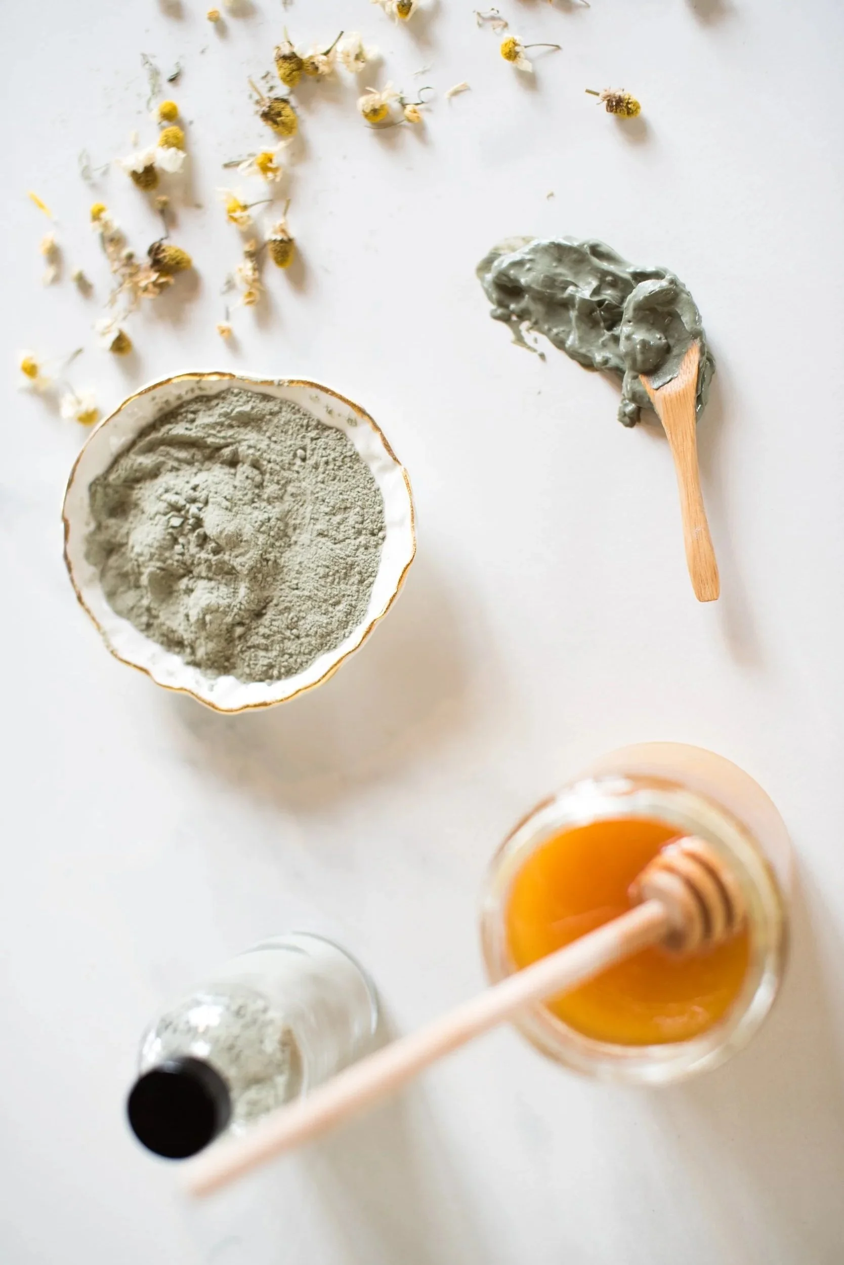 Dried chamomile flowers, a bowl of green clay powder, a wooden spatula with green clay, a glass jar of honey with a honey dipper, and a small jar of powder on a white surface.