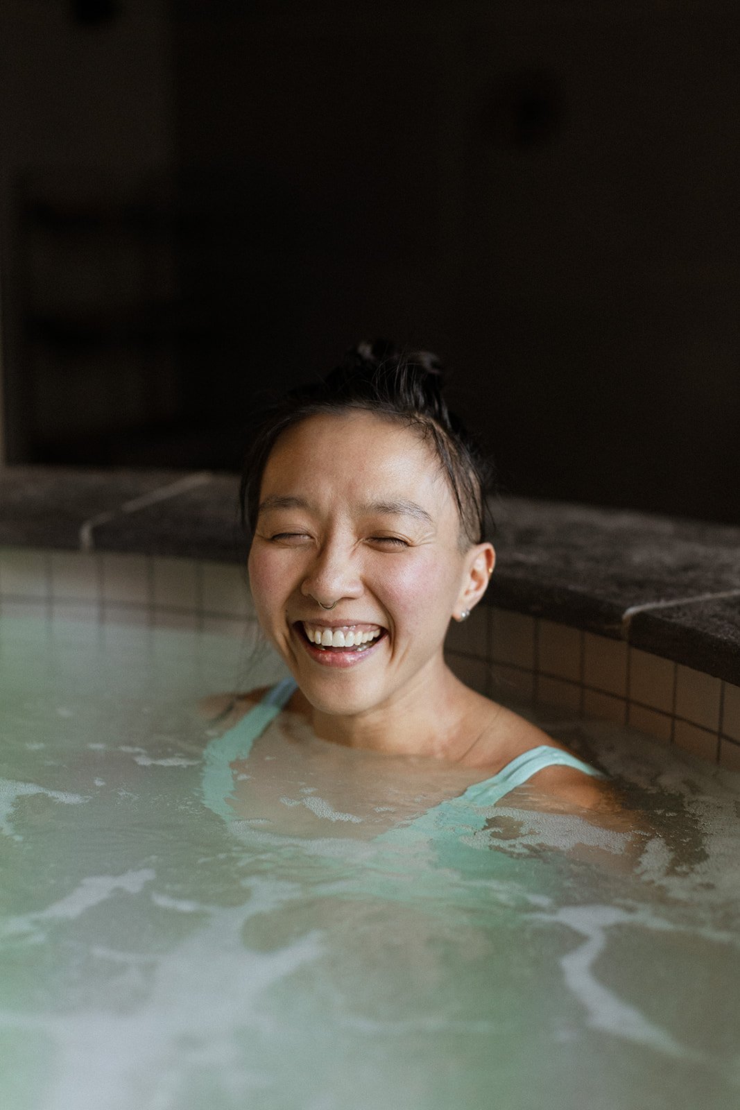 Woman with short dark hair and a septum piercing smiling in a hot tub with dark background.