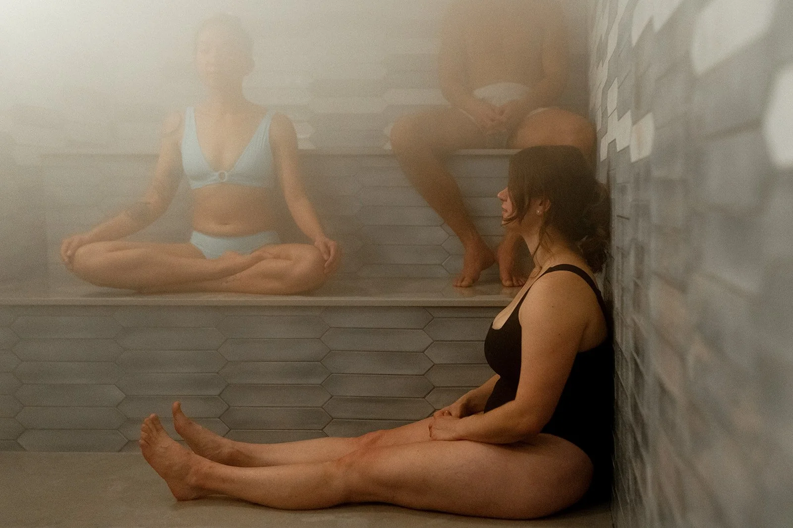 A woman with dark hair sitting on the floor in a sauna, wearing a black swimsuit, with her back against a white brick wall, and two people sitting on a bench inside the sauna, one in a blue swimsuit and the other in possibly underwear, partially obscured and reflected in the steam.