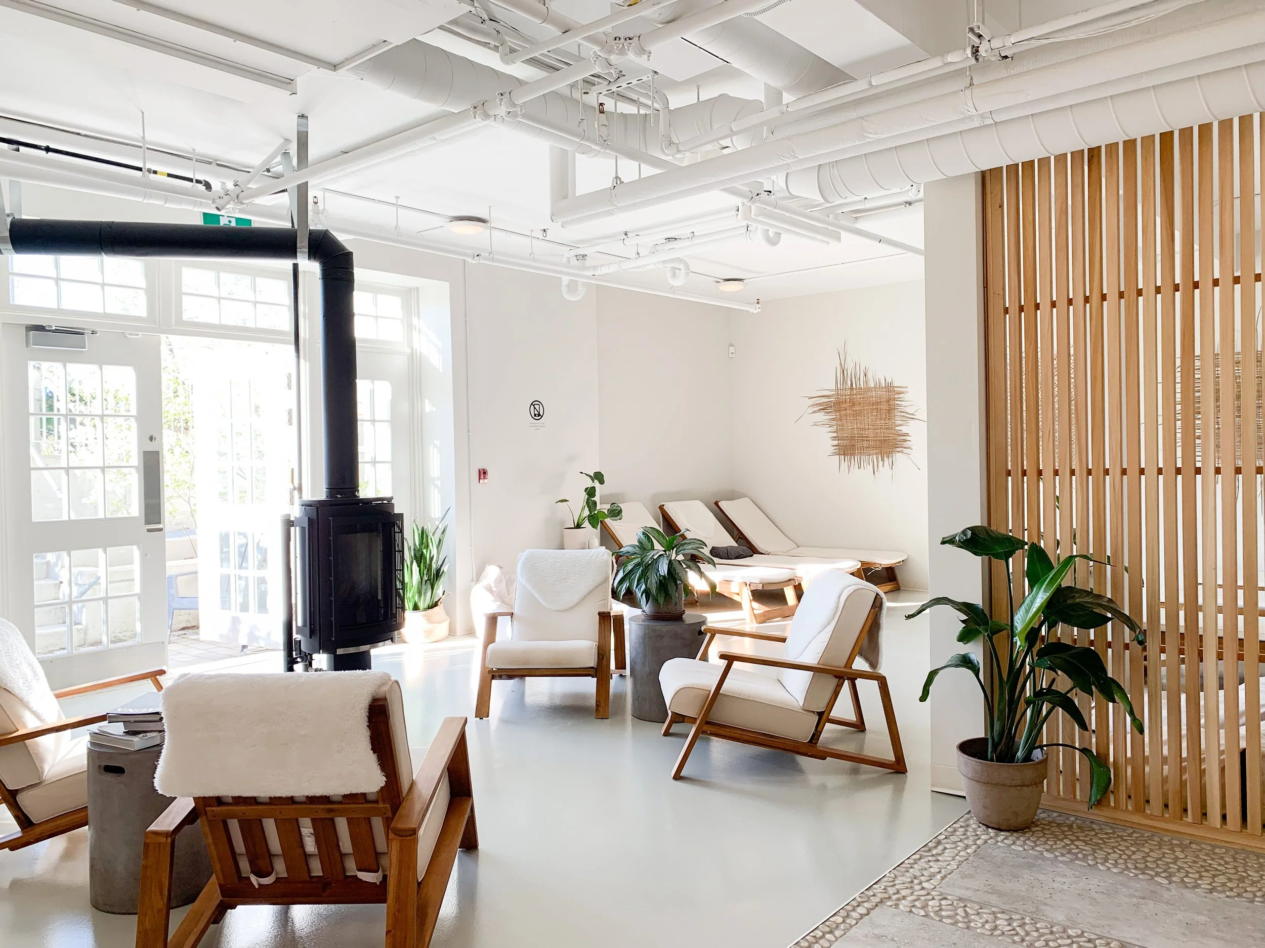 Bright, modern lounge area with wooden chairs, potted plants, a wood stove, and lounge chairs near the window, decorated with minimalist art and natural accents.