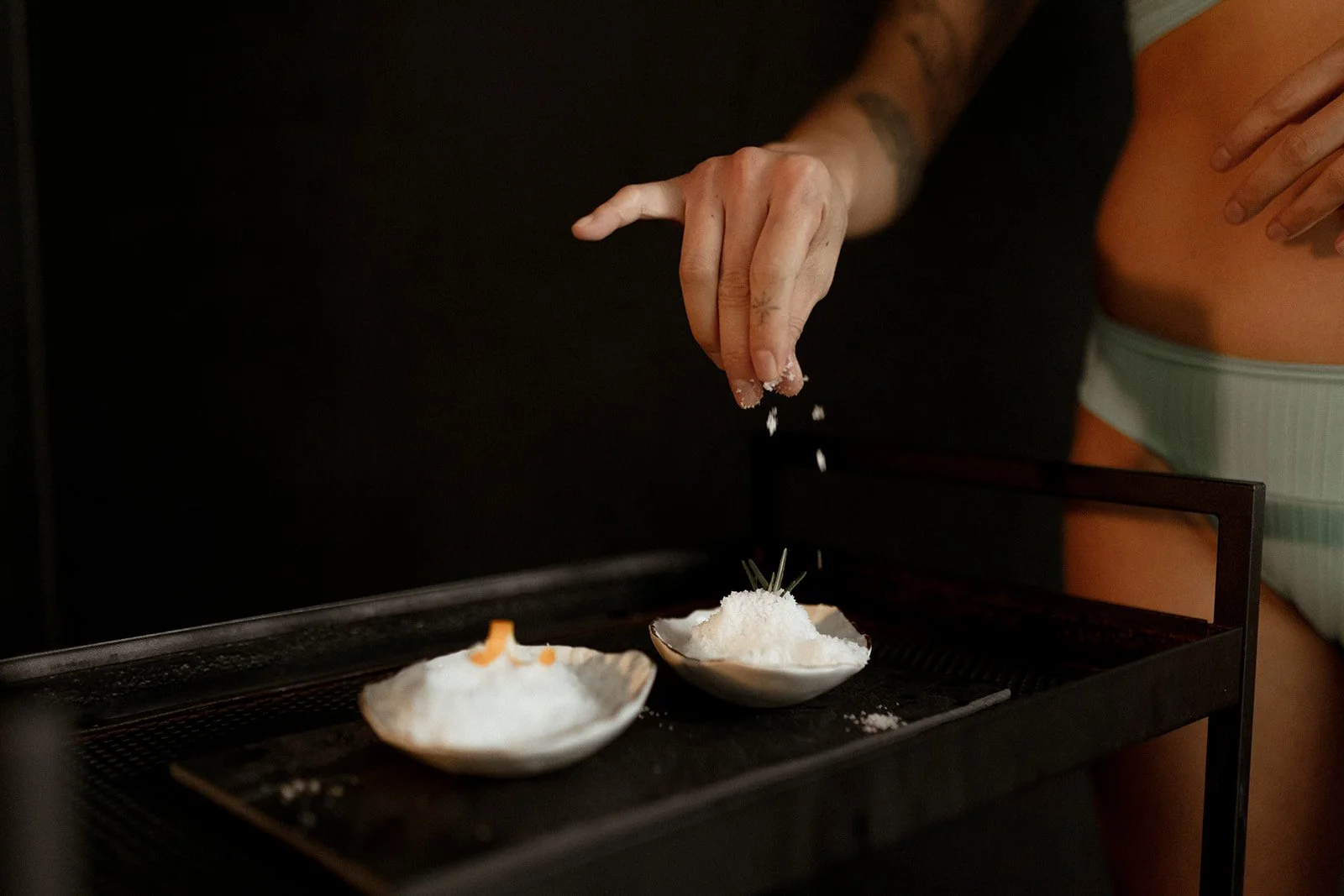Person sprinkling salt onto two small white shell-shaped dishes on a tray.
