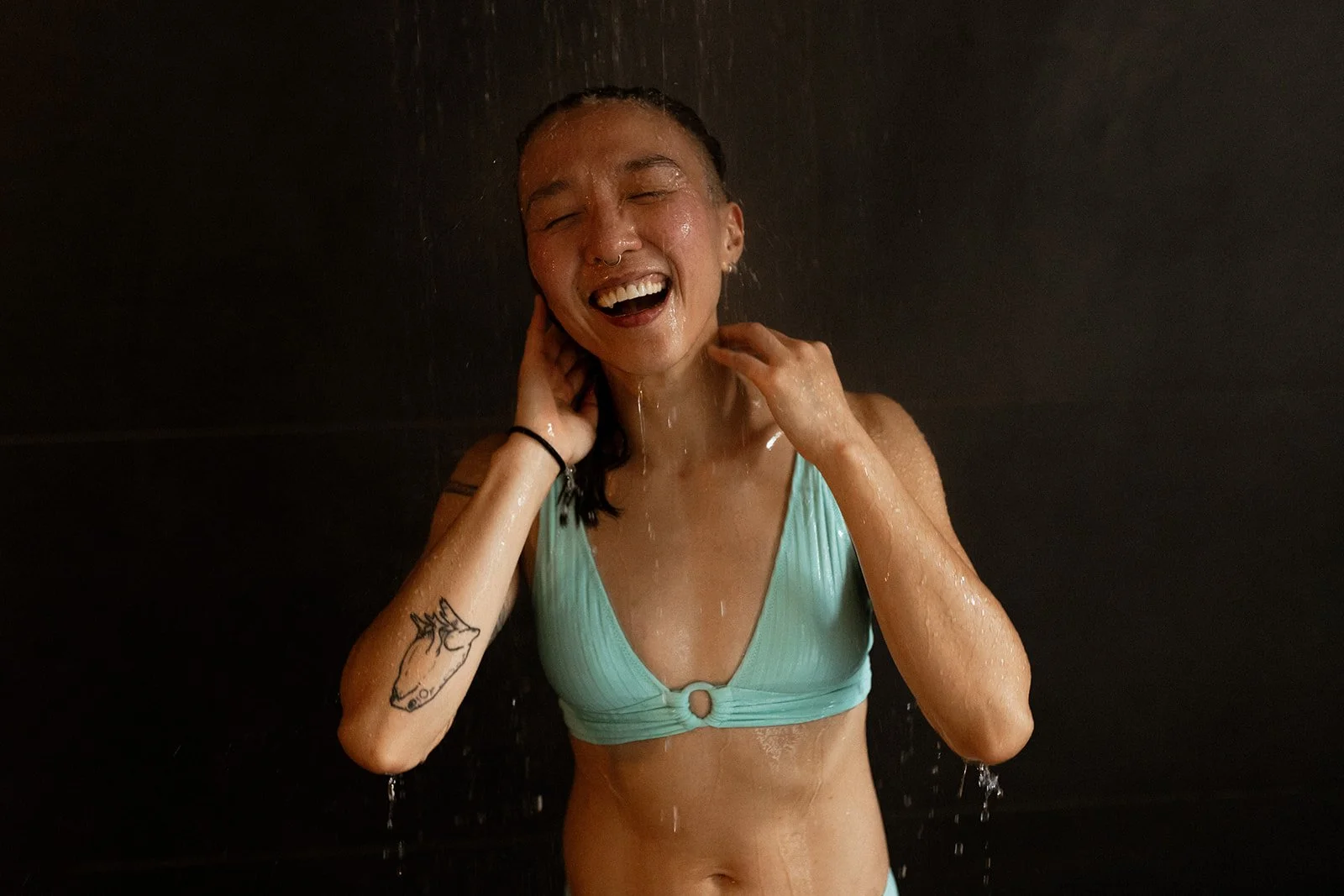 A woman enjoying a shower, smiling with eyes closed, wearing a light blue bathing suit, with water running down her face and body, in a dark shower area.