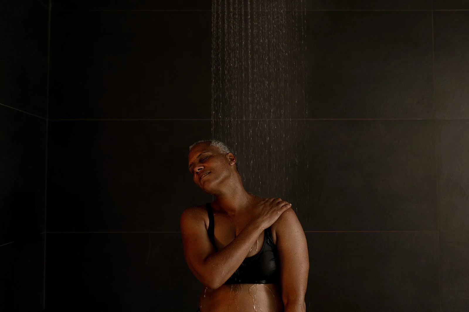 A person in a black tank top stands under a shower with water falling down, with eyes closed and a peaceful expression.