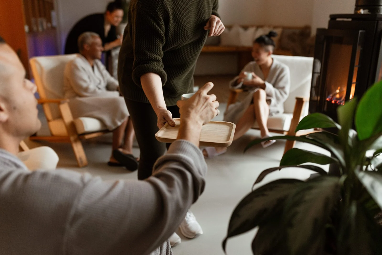 People sitting in a cozy living room, with a woman serving tea from a tray to a young man, while others relax on chairs and couches in the background.