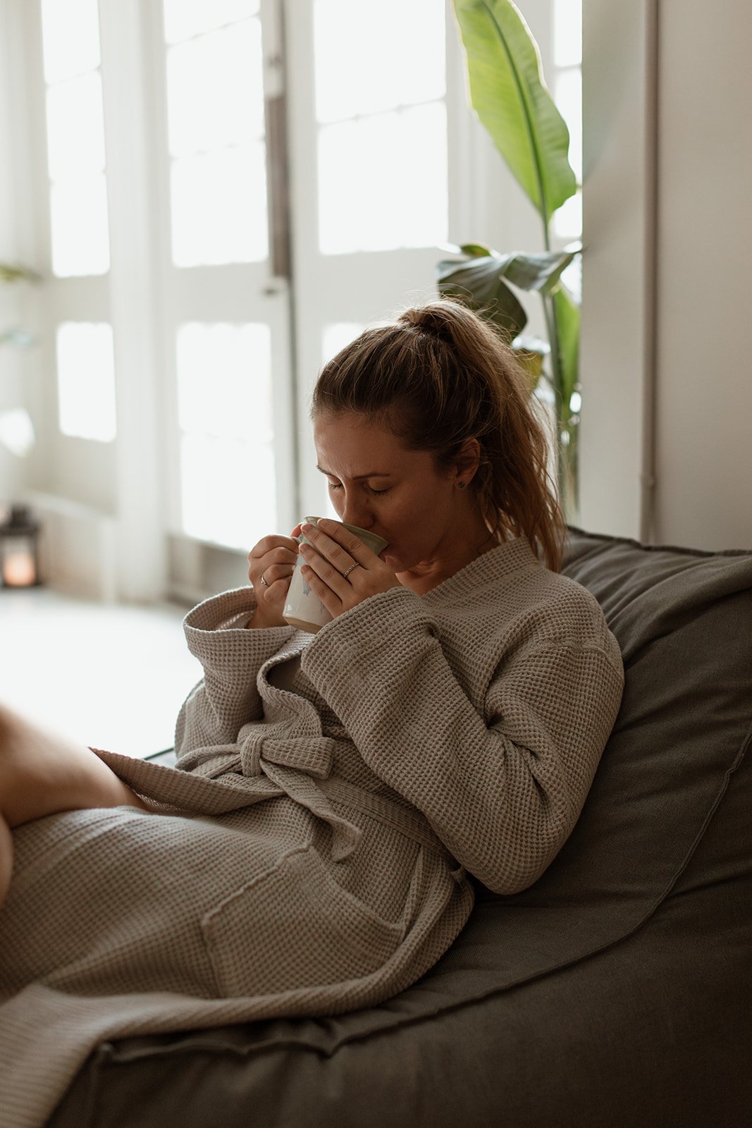 Woman sitting on a sofa, wearing a beige robe, drinking from a mug inside a cozy home with sunlight coming through a glass door.