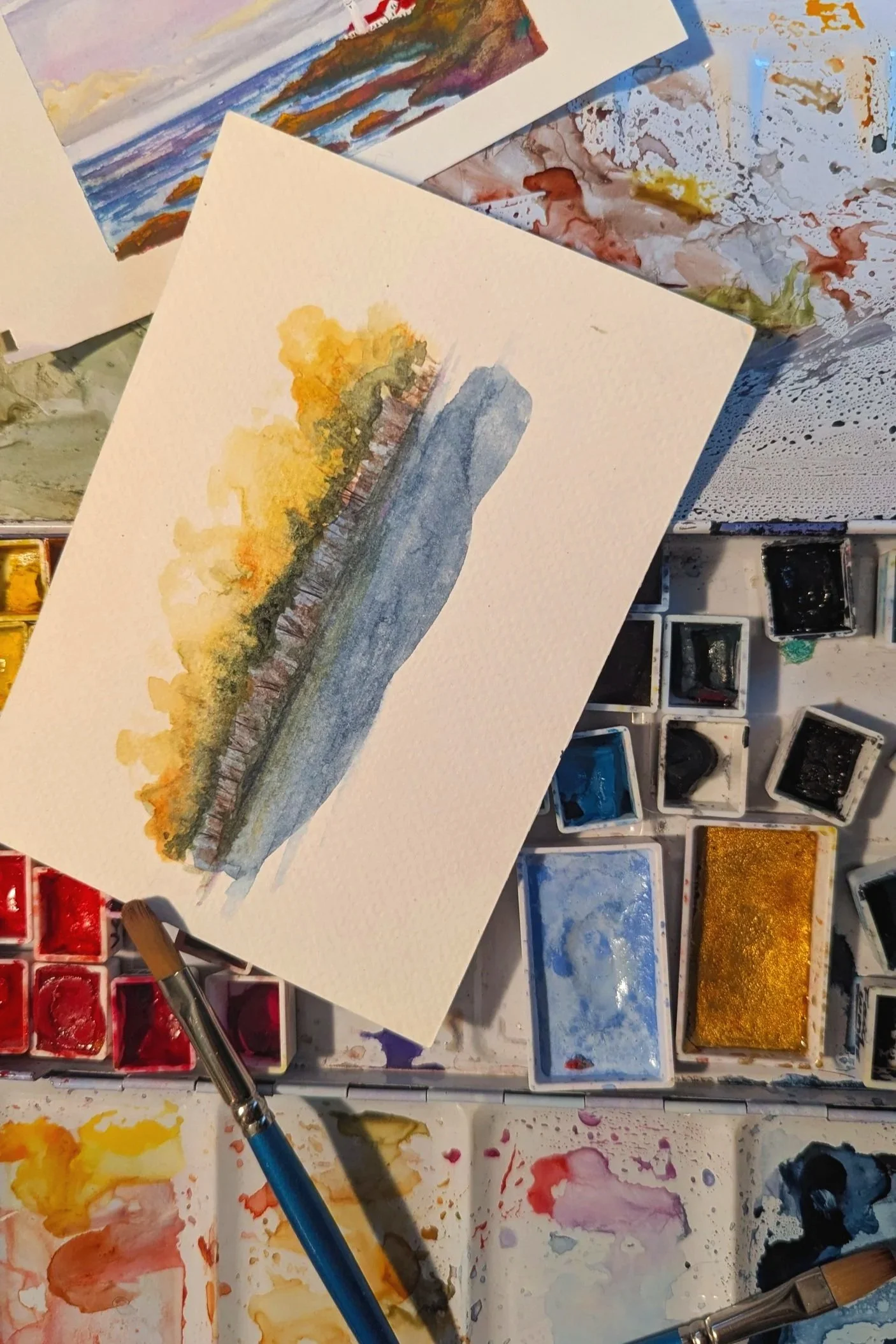 A watercolor painting of a landscape with a body of water, trees, and a cloudy sky, surrounded by watercolor paints and brushes.