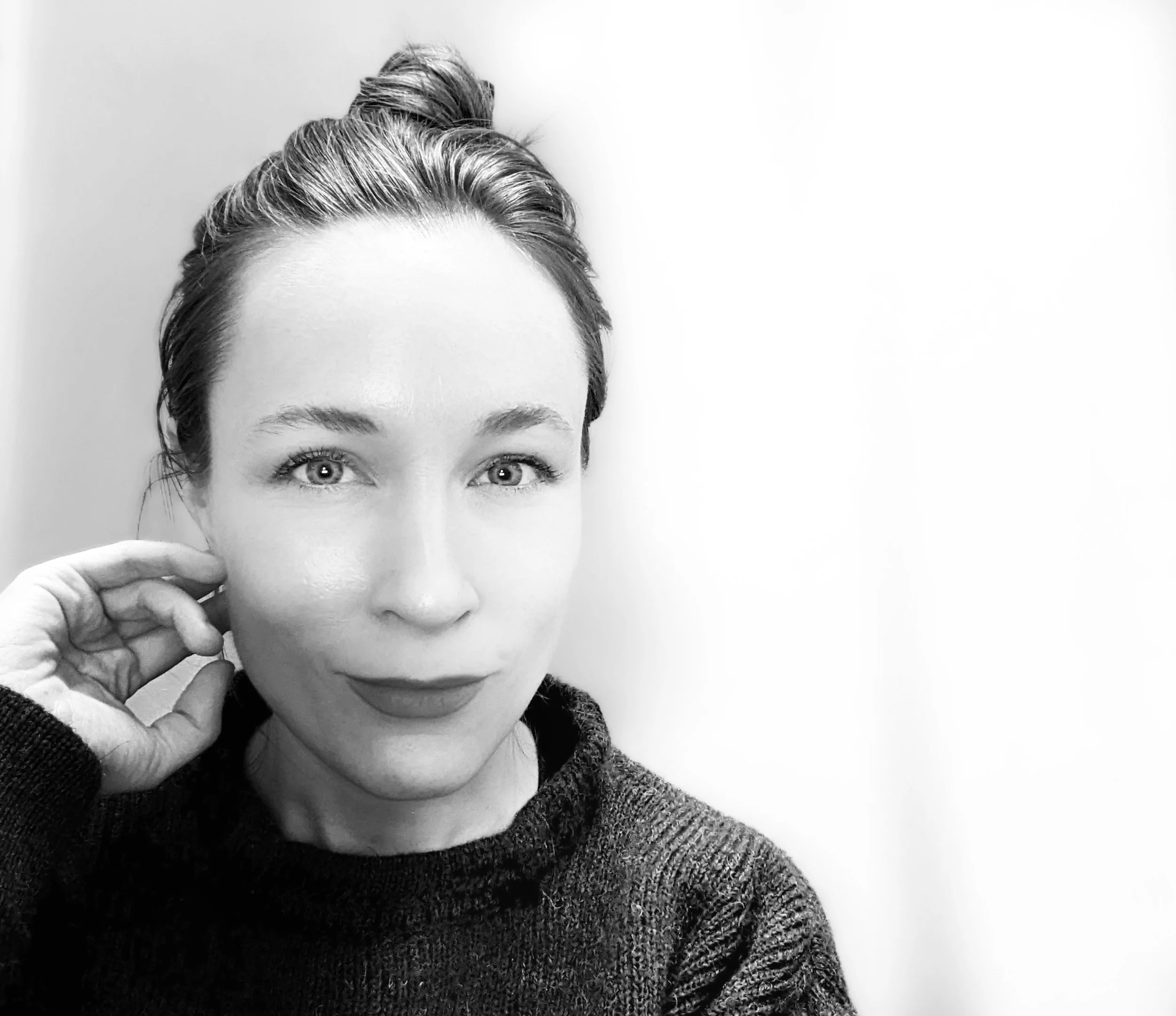 Black and white portrait of a woman with light eyes, wearing a dark sweater, touching her face with her left hand and smiling slightly.