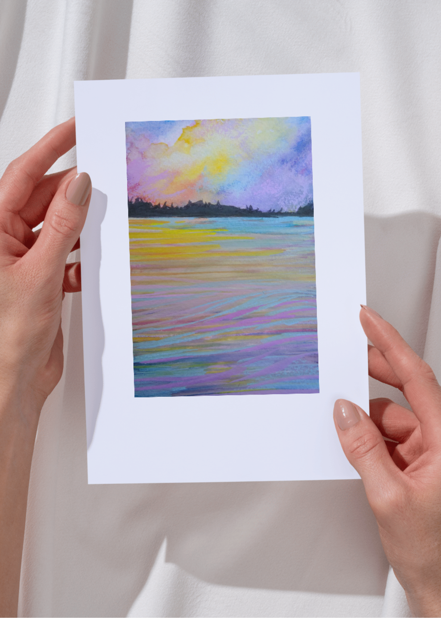 Person holding a watercolor painting of a sunset over a body of water with colorful sky and reflections.