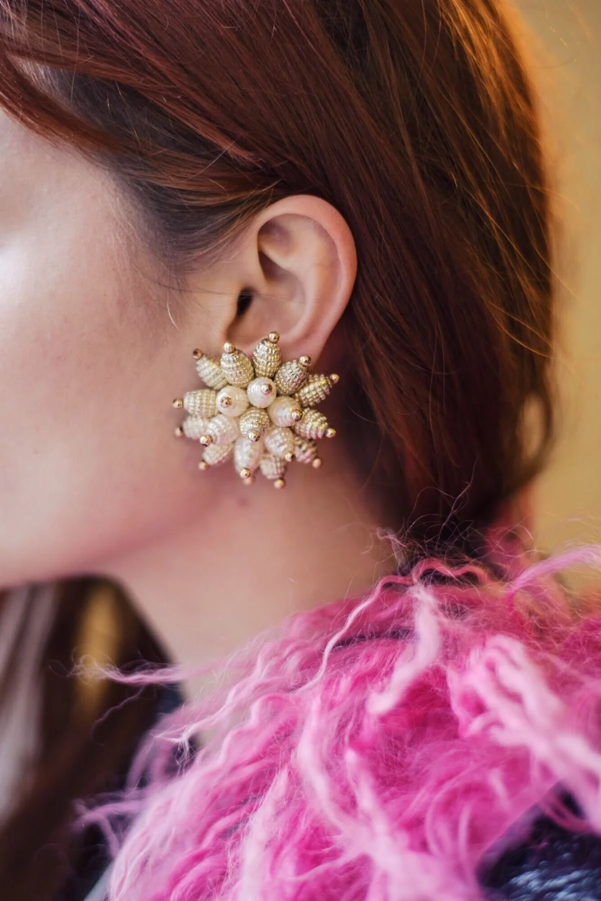Close-up of a woman's ear adorned with a large vintage gold beaded statement earring, styled with a pink fluffy textured coat.
