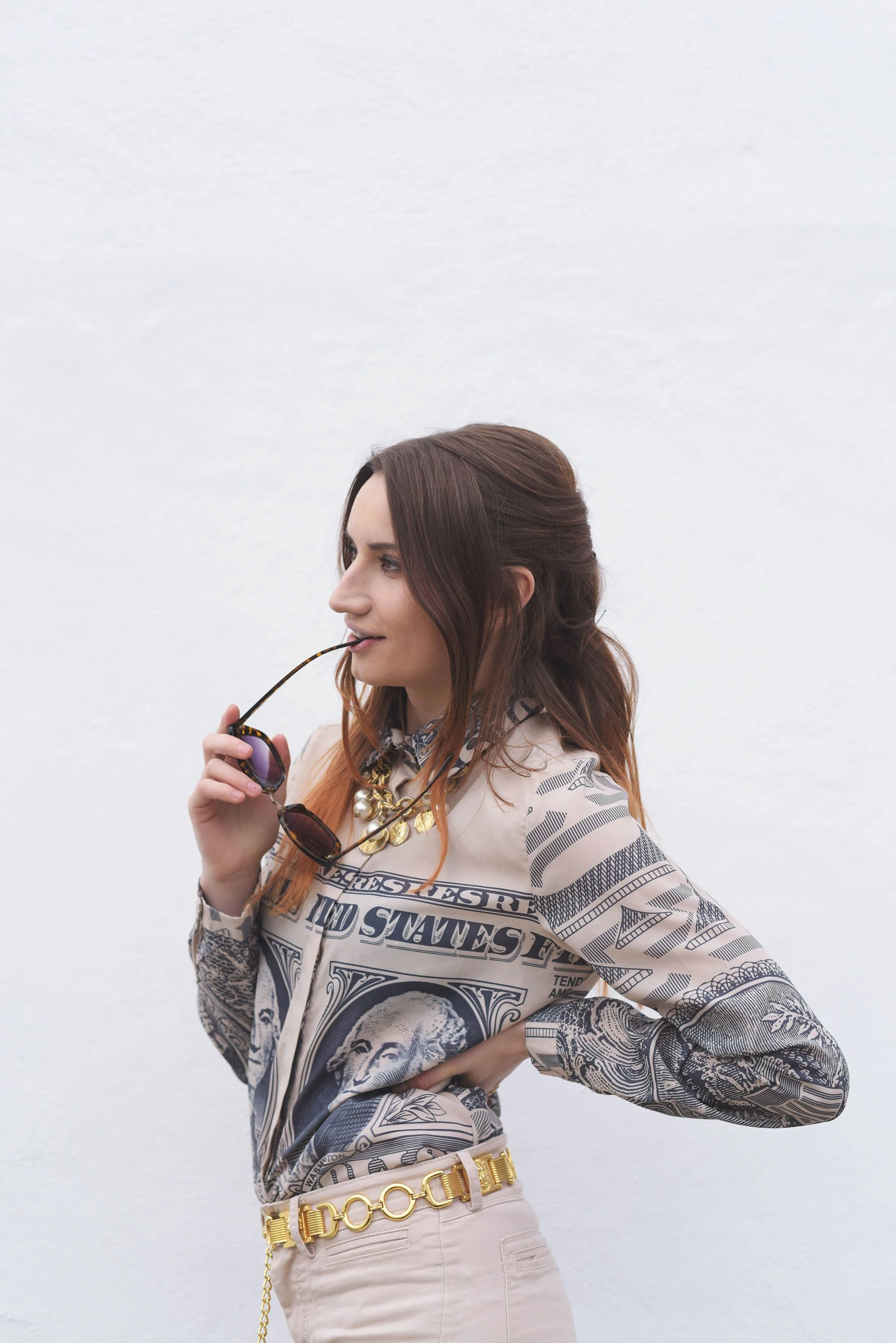 Close-up of a woman in a vintage money-print shirt with a chunky gold chain belt and layered necklaces, holding sunglasses near her lips, posing against a white wall.