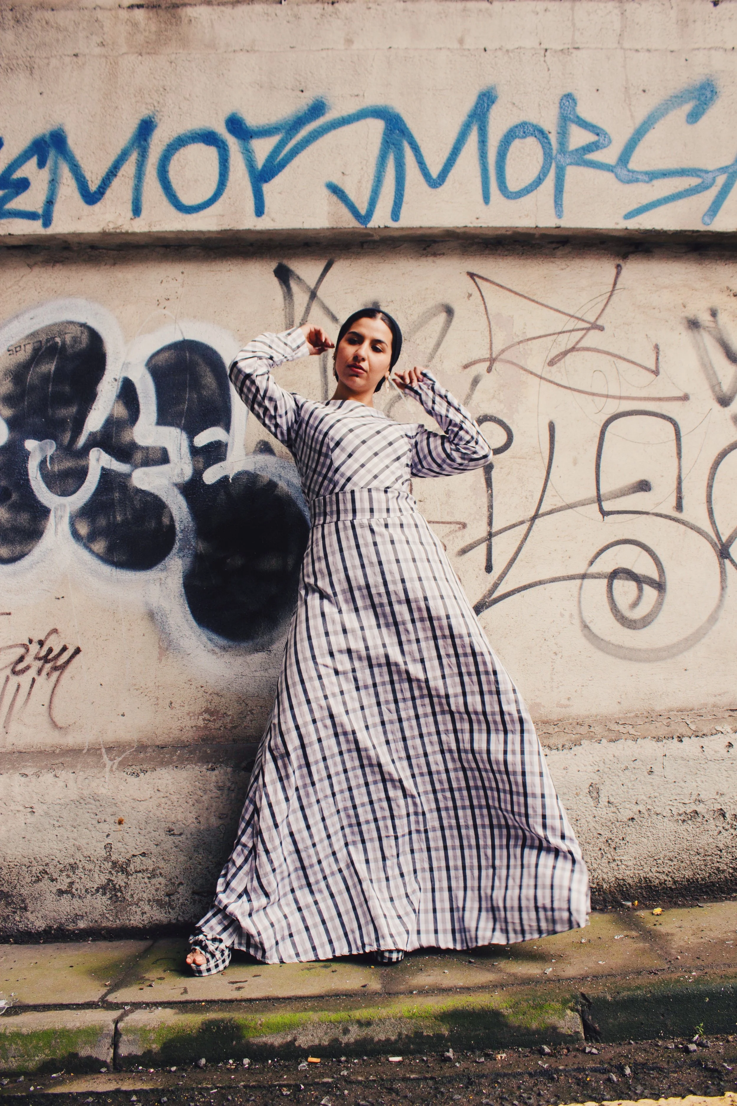Woman in a checkered maxi dress posing against a graffiti-covered urban wall, combining elegance with street style.