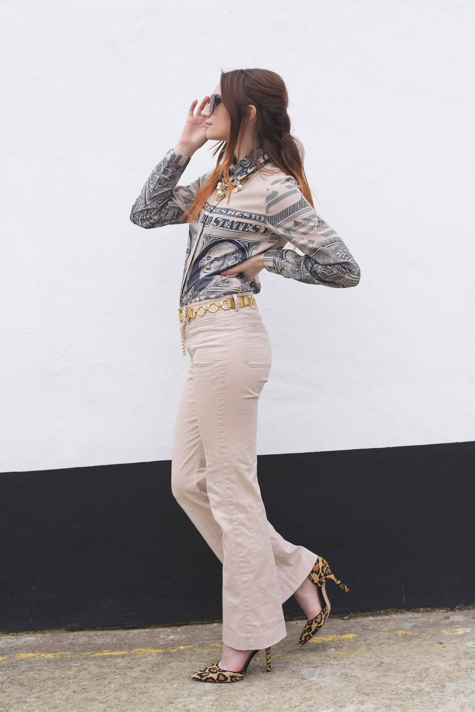 Woman wearing a money print shirt, beige flared pants, gold belt, and leopard print heels, posing against a minimal white and black wall.