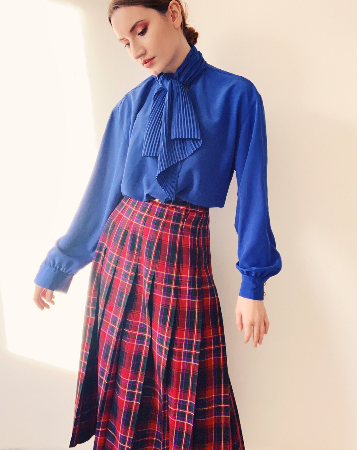 Model wearing a royal blue pleated blouse with a dramatic bow and a high-waisted vintage tartan skirt, styled and photographed by Louisa Rogers in a sunlit studio space.