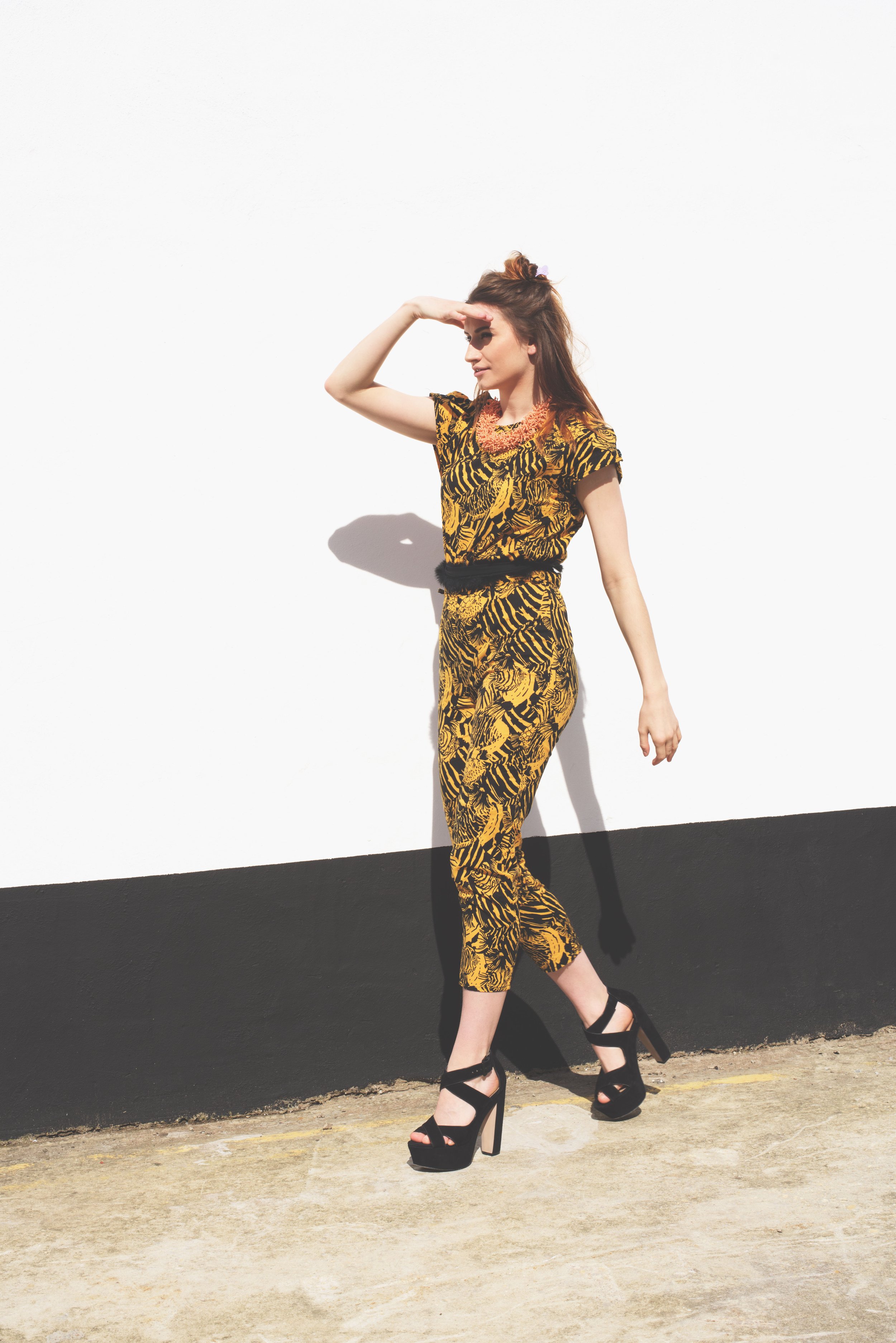 Louisa Rogers modeling a bold yellow and black printed jumpsuit with platform heels, styled and photographed against a minimalist white and black urban wall in bright sunlight
