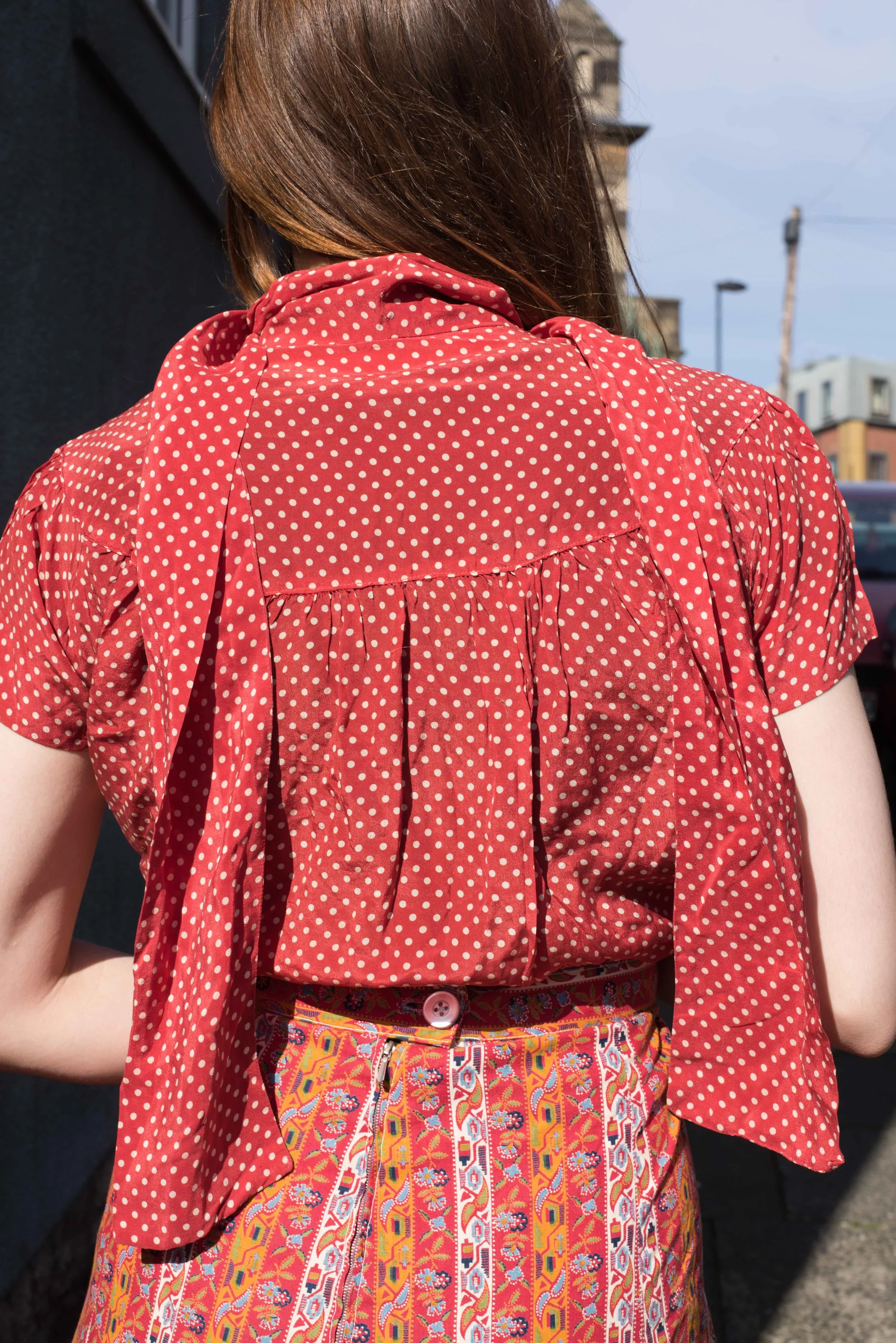 Back view of a woman wearing a red polka dot blouse paired with a colorful patterned skirt, photographed in natural sunlight on an urban street.