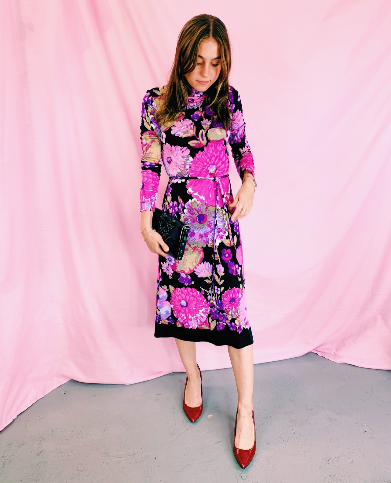 Woman wearing a retro floral dress with pink and purple blooms on a black background, styled with red heels and a black clutch, standing in front of a soft pink backdrop.