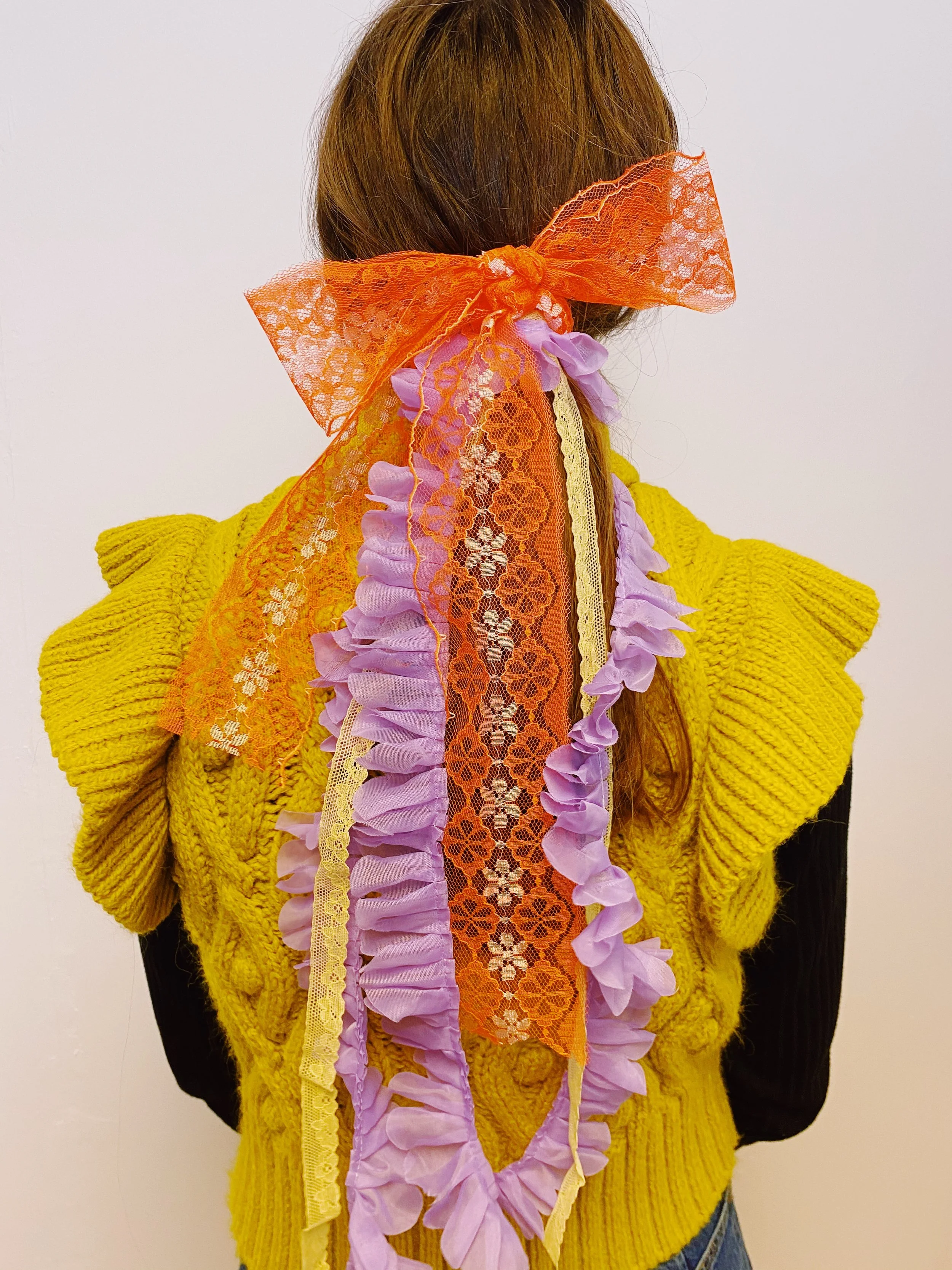 Back view of a hairstyle adorned with vintage ribbons, including an orange lace bow and cascading purple and yellow ruffled trims, styled by Louisa Rogers and worn over a textured mustard-yellow knit top.