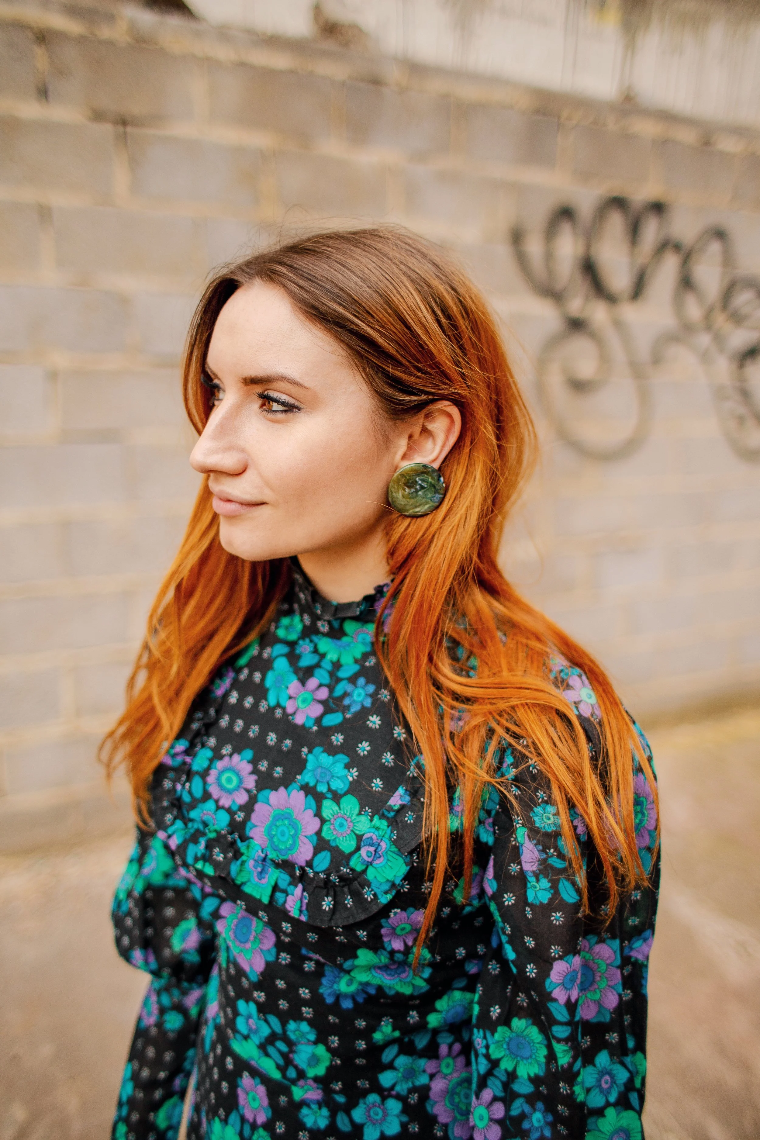 Portrait of a woman with long red hair wearing a vintage-inspired floral dress and large green earrings, standing in front of a weathered brick wall with graffiti.