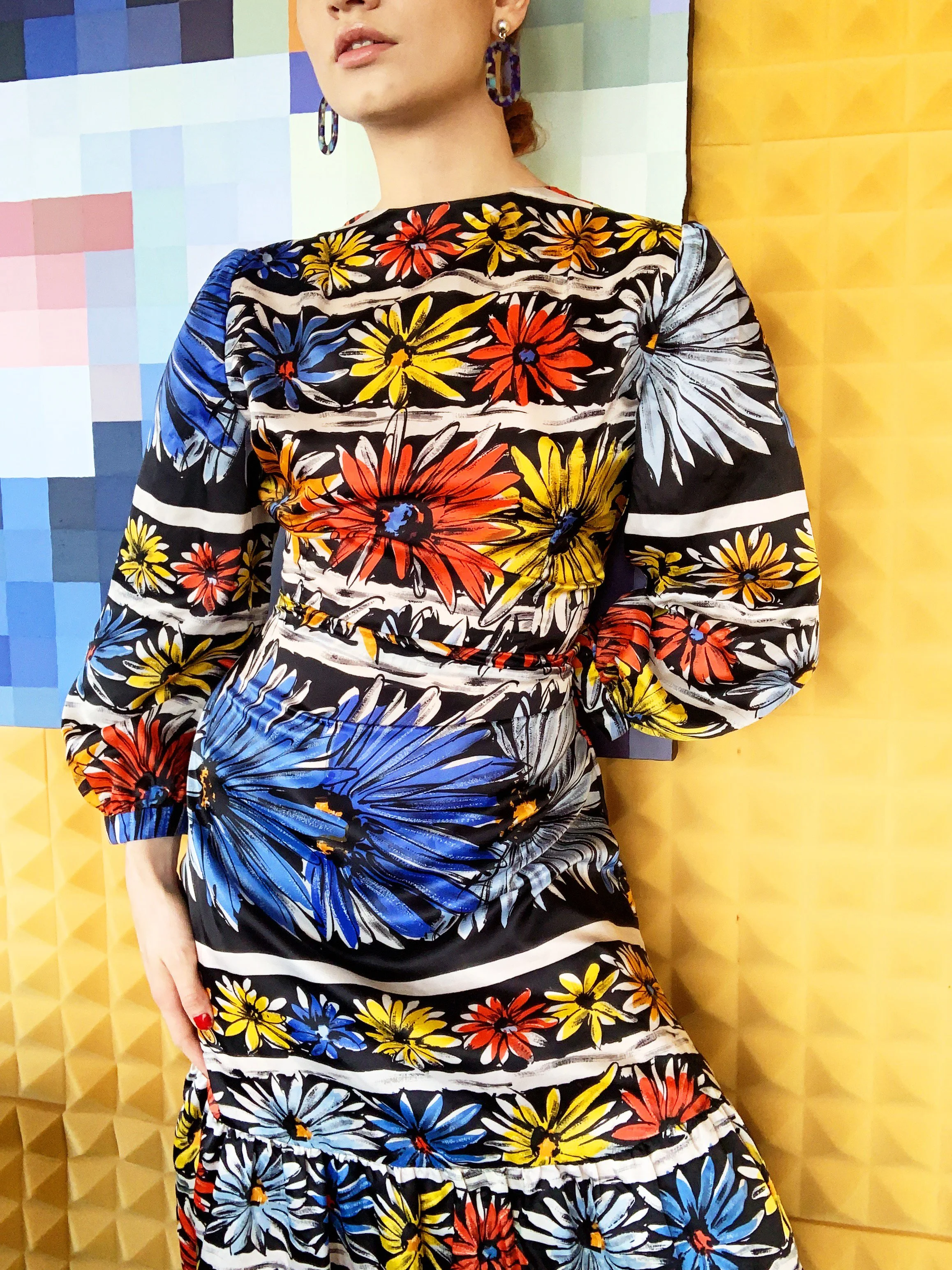 Woman wearing a bold floral print dress featuring red, yellow, blue, and white flowers against a black background, posing in front of a vibrant geometric wall with blue and yellow tones.