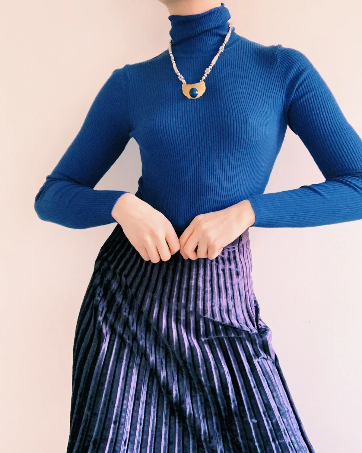 Close-up of a model wearing a cobalt blue ribbed turtleneck paired with a pleated purple velvet skirt and a sculptural gold and stone necklace, styled and photographed by Louisa Rogers.