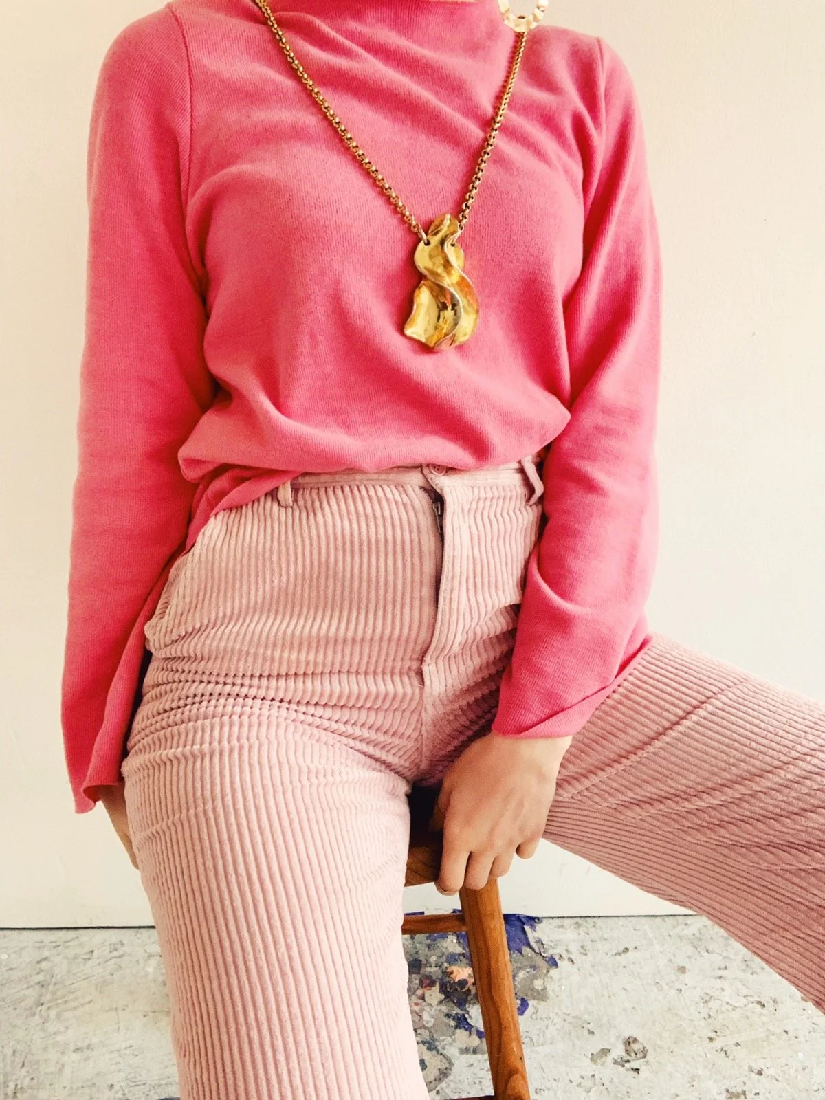 Close-up of a monochrome outfit featuring a pink turtleneck and blush corduroy trousers, styled with a bold sculptural gold necklace, photographed by Louisa Rogers in a studio setting.