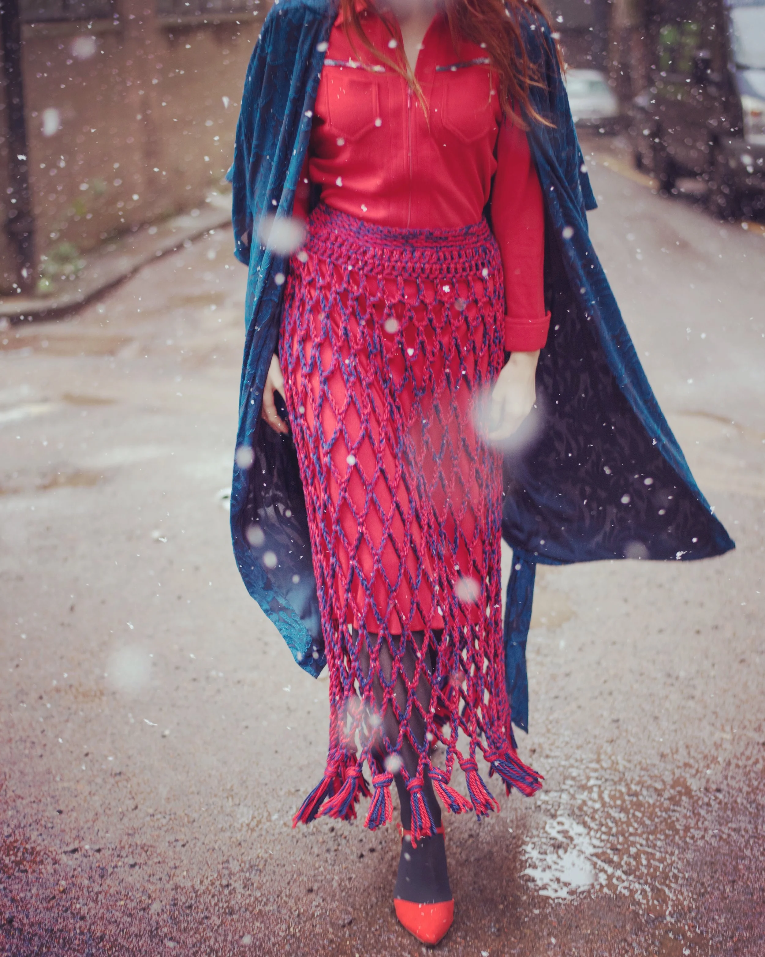 Fashion portrait of a woman walking through falling snow in a bold red macrame skirt, red shirt, and teal velvet coat, styled and photographed by Louisa Rogers.