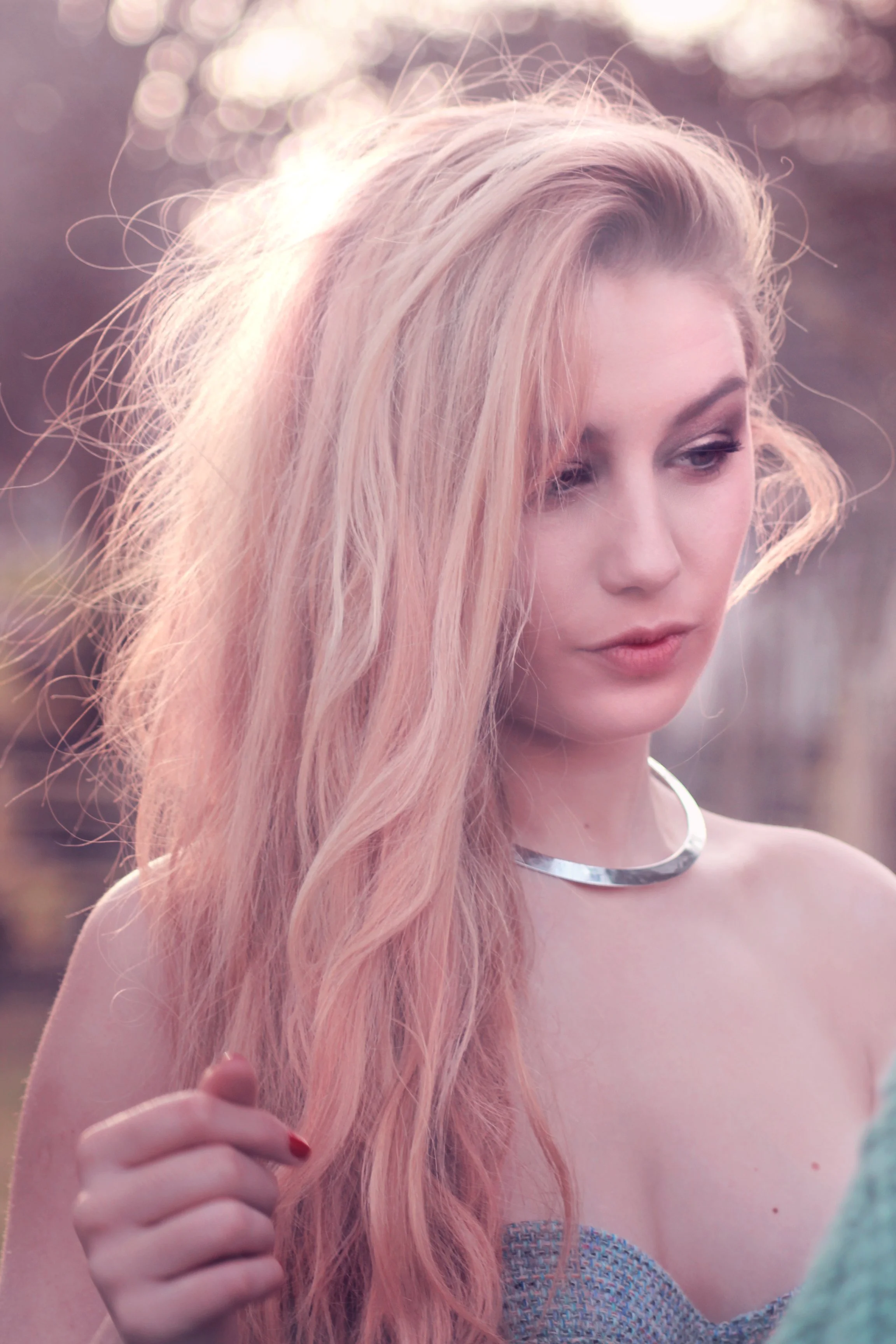 Soft focus outdoor beauty portrait of a blonde woman with windswept hair looking downward, wearing a silver torque collar necklace and a blue strapless bralette, with a warm pink-toned bokeh background