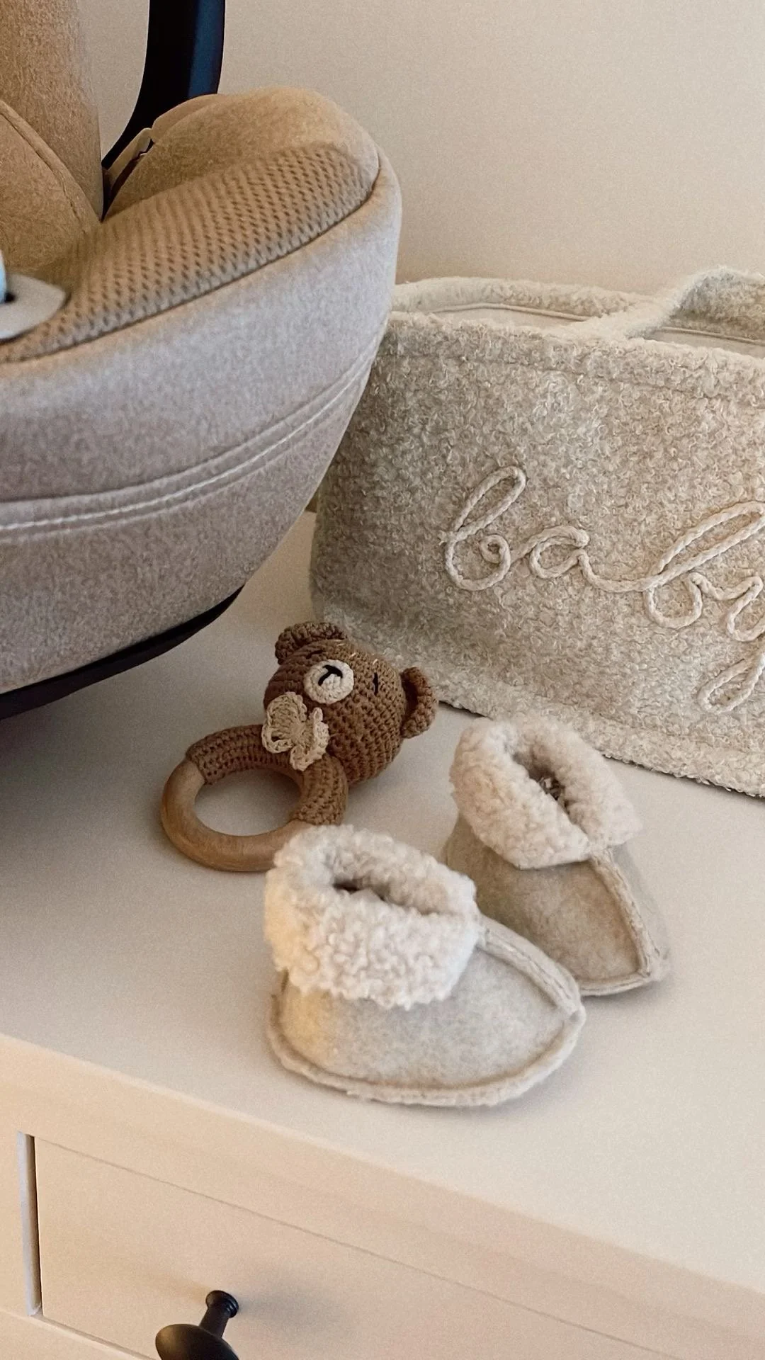 A pair of gray baby booties with fluffy trim, a beige plush rattle with a bear face, a padded storage box with the word "baby" embroidered, and part of a beige upholstered chair on a white surface.