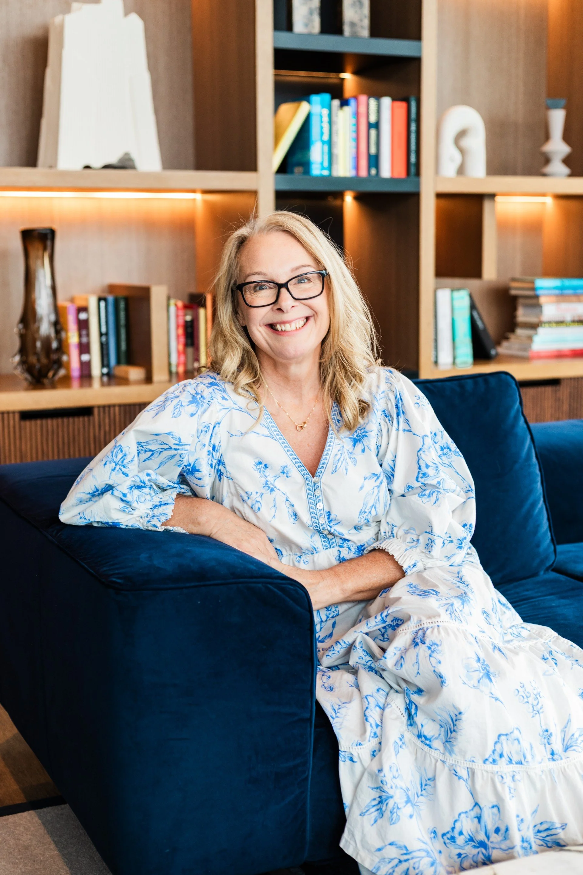 A woman with glasses and blonde hair smiling while sitting on a dark blue velvet sofa in a well-decorated living room with wooden shelves filled with books and decorative objects in the background.