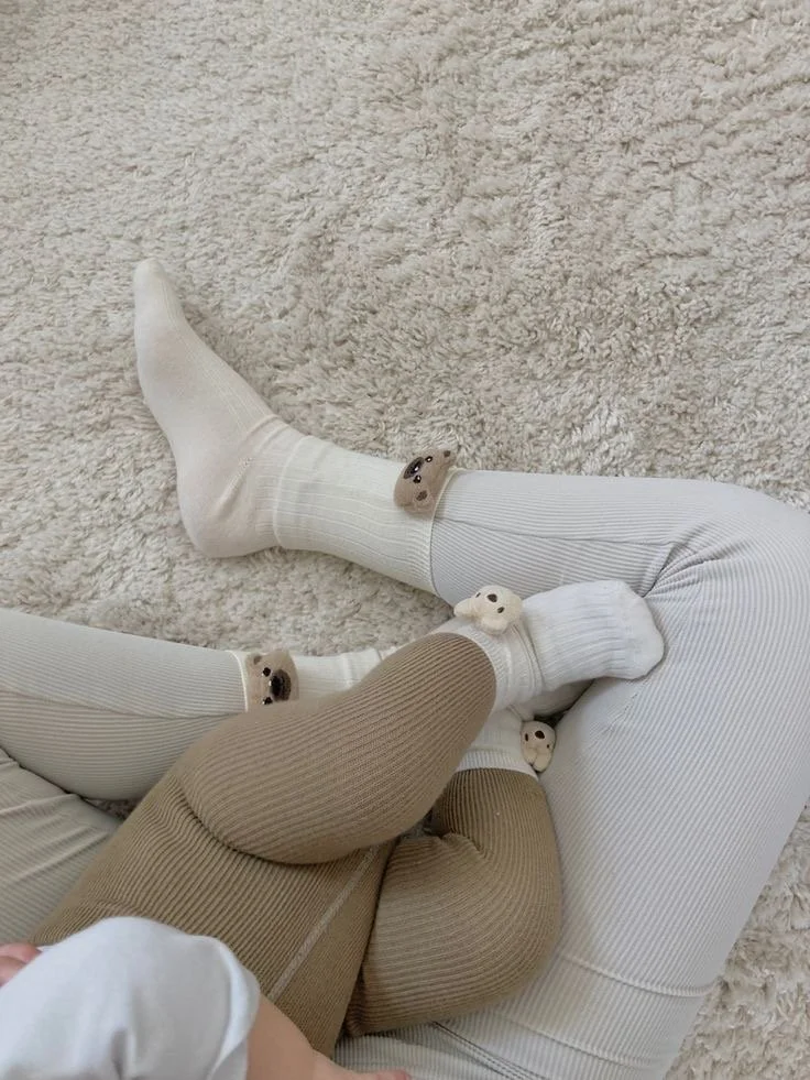 Close-up of legs and feet of two people sitting on a plush beige carpet, wearing matching fuzzy bear paw slippers and cozy clothing in neutral tones.