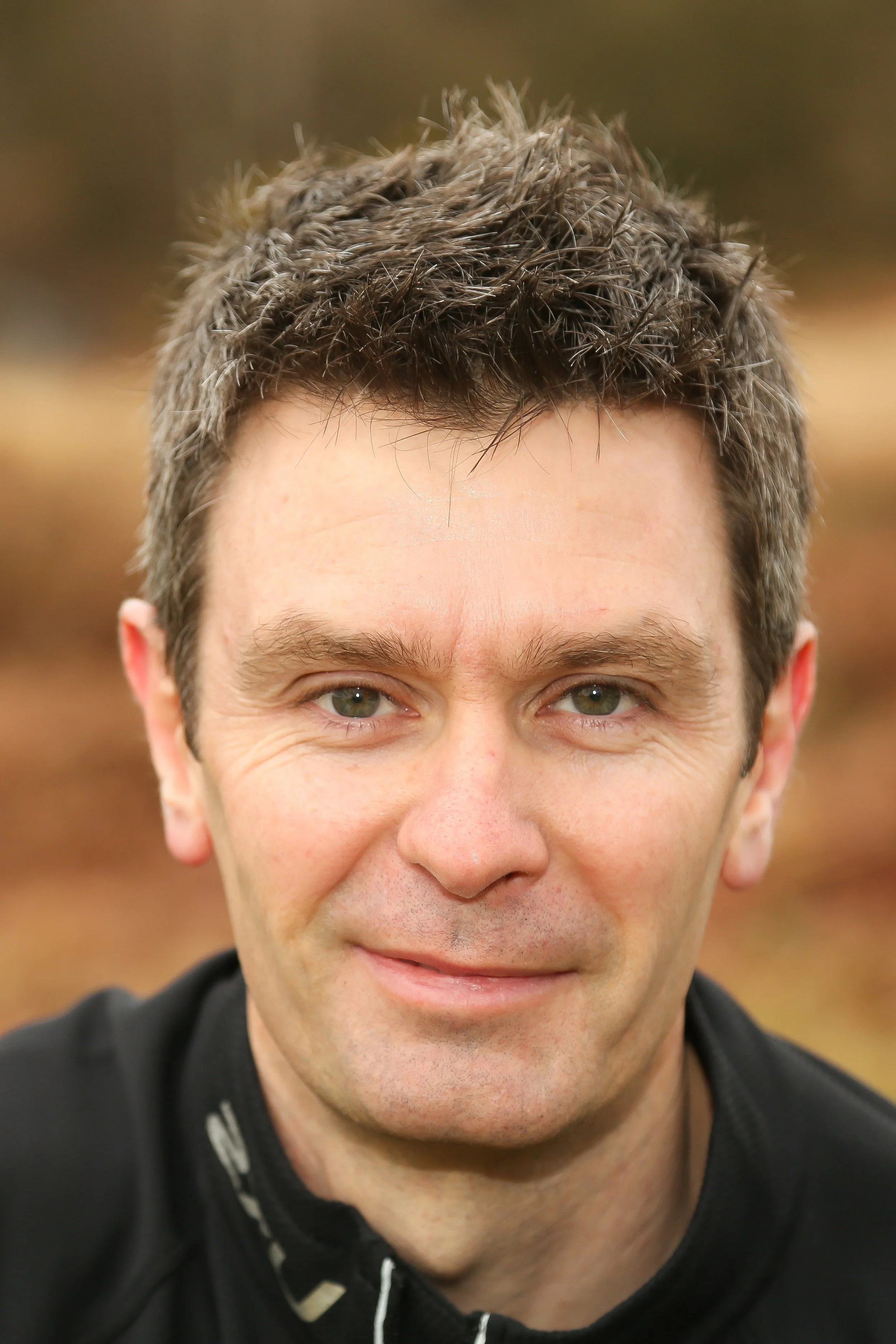 Close-up portrait of a man with short, dark, slightly curly hair and blue eyes, smiling slightly, outdoors with autumn-colored blurred background.