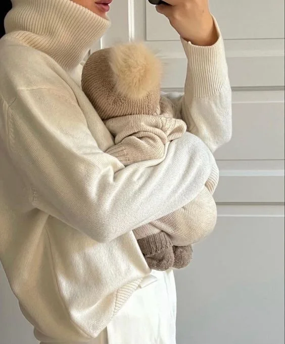 A person holding a bundled-up baby, both wearing beige and cream-colored knit clothing, standing in front of a light-colored wall.
