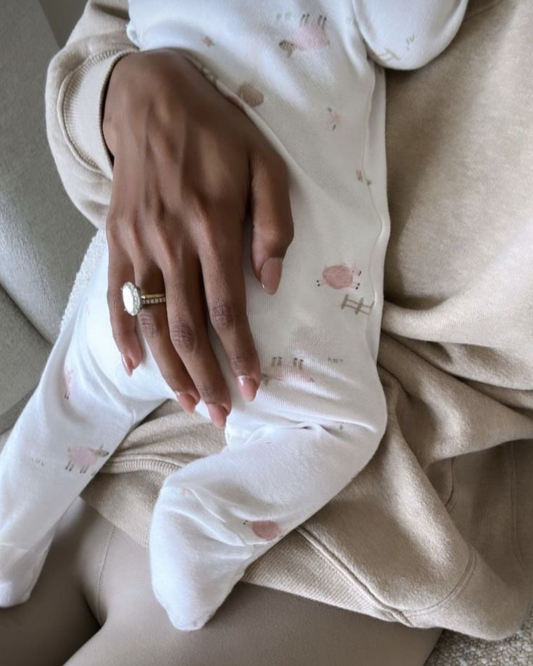 A close-up of a person’s hand resting on their lap, wearing a large ring with a white stone, sitting on a beige chair, wearing a light-colored sweatshirt and white pants with pink spots.