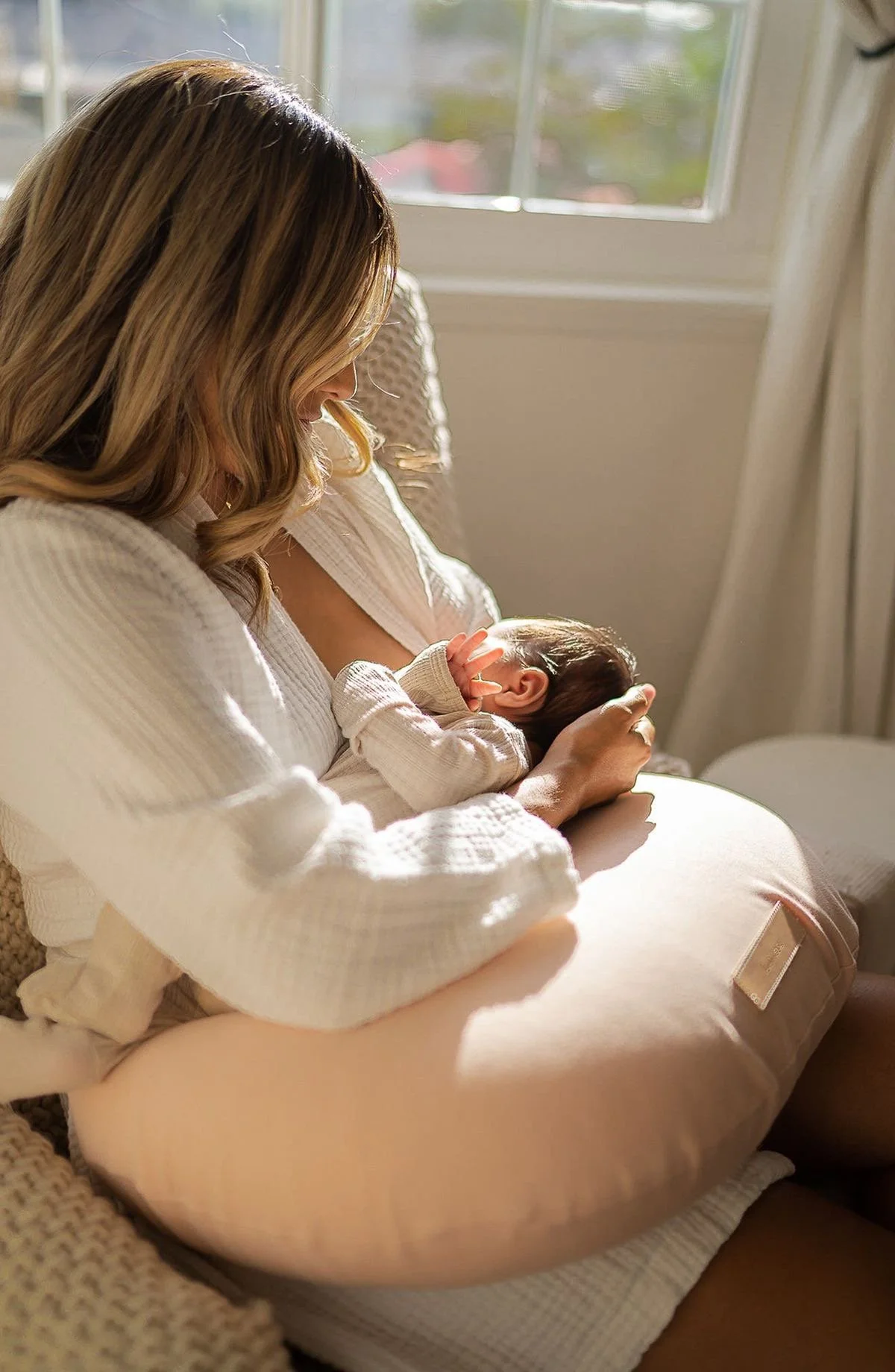A woman breastfeeding a baby in a cozy room with sunlight coming through a window.