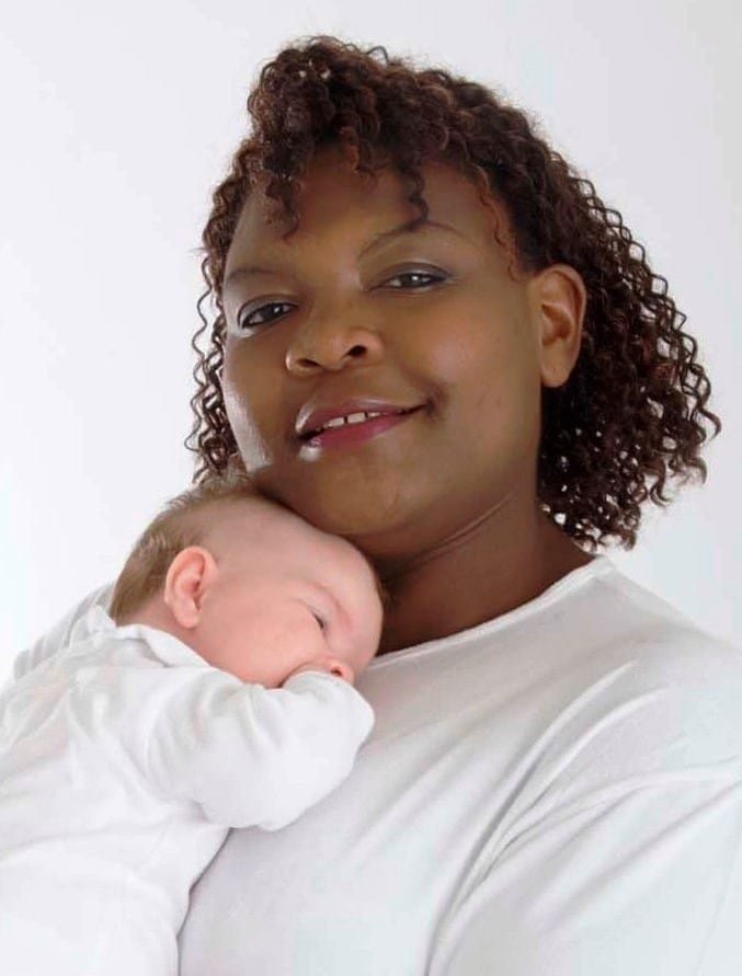 A woman holding a sleeping baby on her shoulder, both wearing white shirts, against a plain light background.