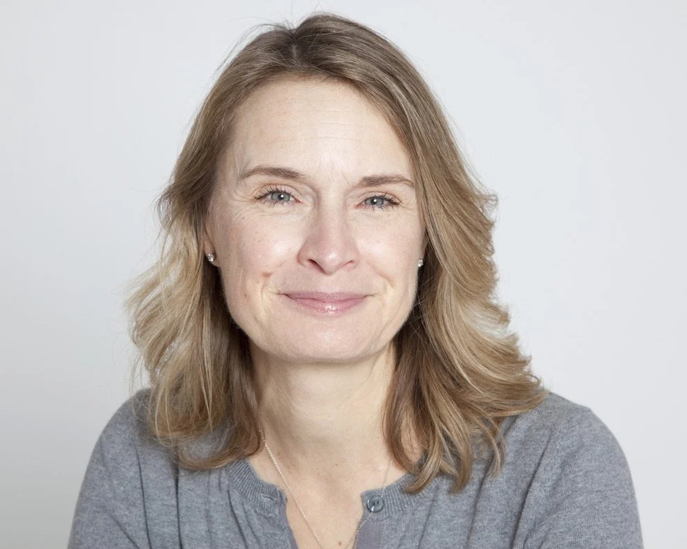 A woman with light skin, shoulder-length wavy light brown hair, and blue eyes smiling in front of a plain light gray background.
