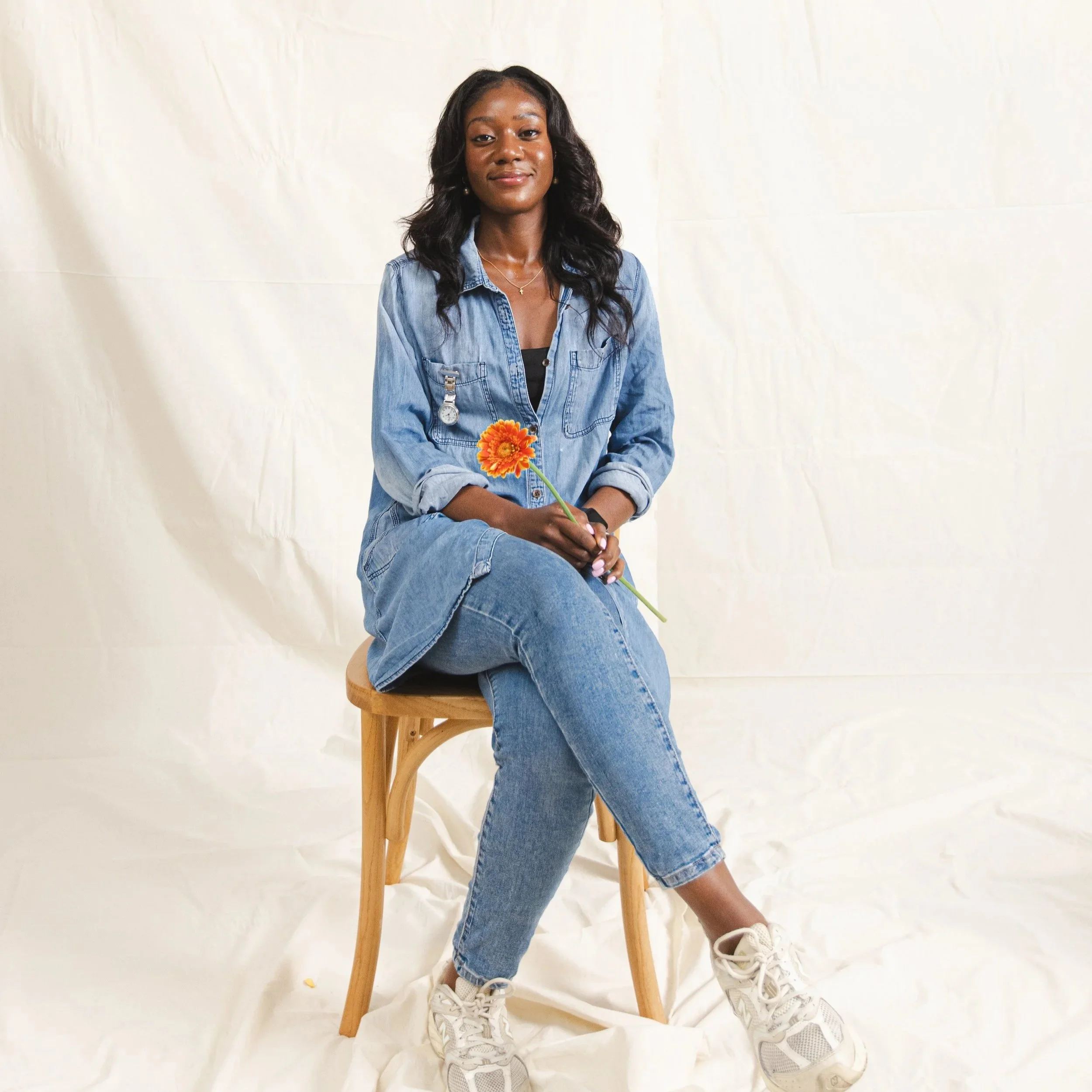 A woman sitting on a modern, off-white textured sofa in a room with white curtains and green plants, wearing a blazer, patterned matching pants and boots, with her hands clasped on her lap.