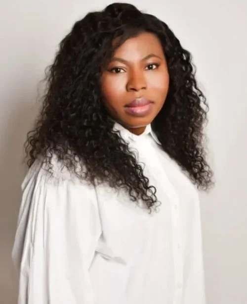 A woman with long, curly black hair wearing a white shirt, standing against a plain background.
