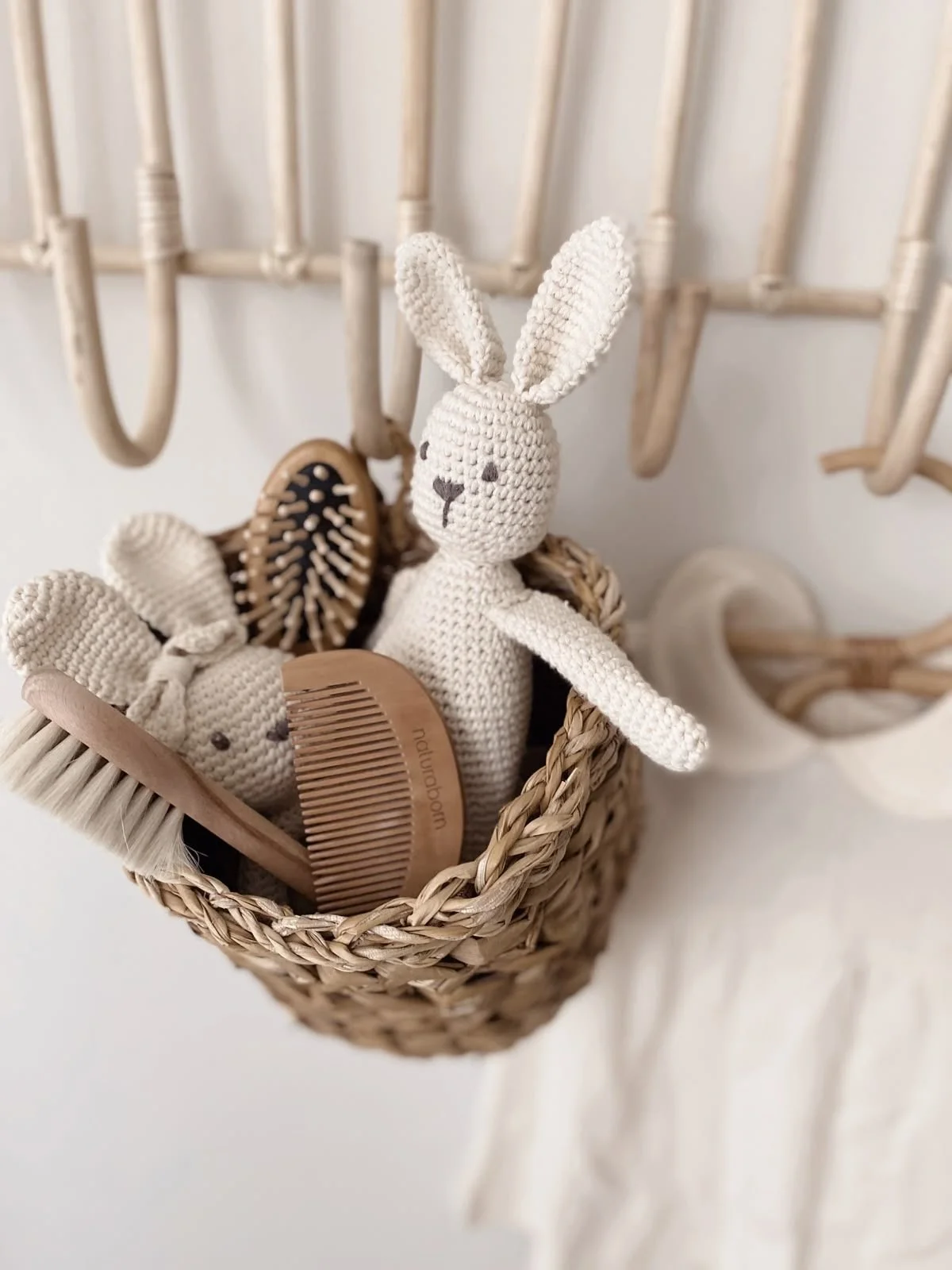 Cream-colored knitted bunny stuffed toy with long ears in a wicker basket, surrounded by a hairbrush and comb, hanging on a wall with wooden hooks.