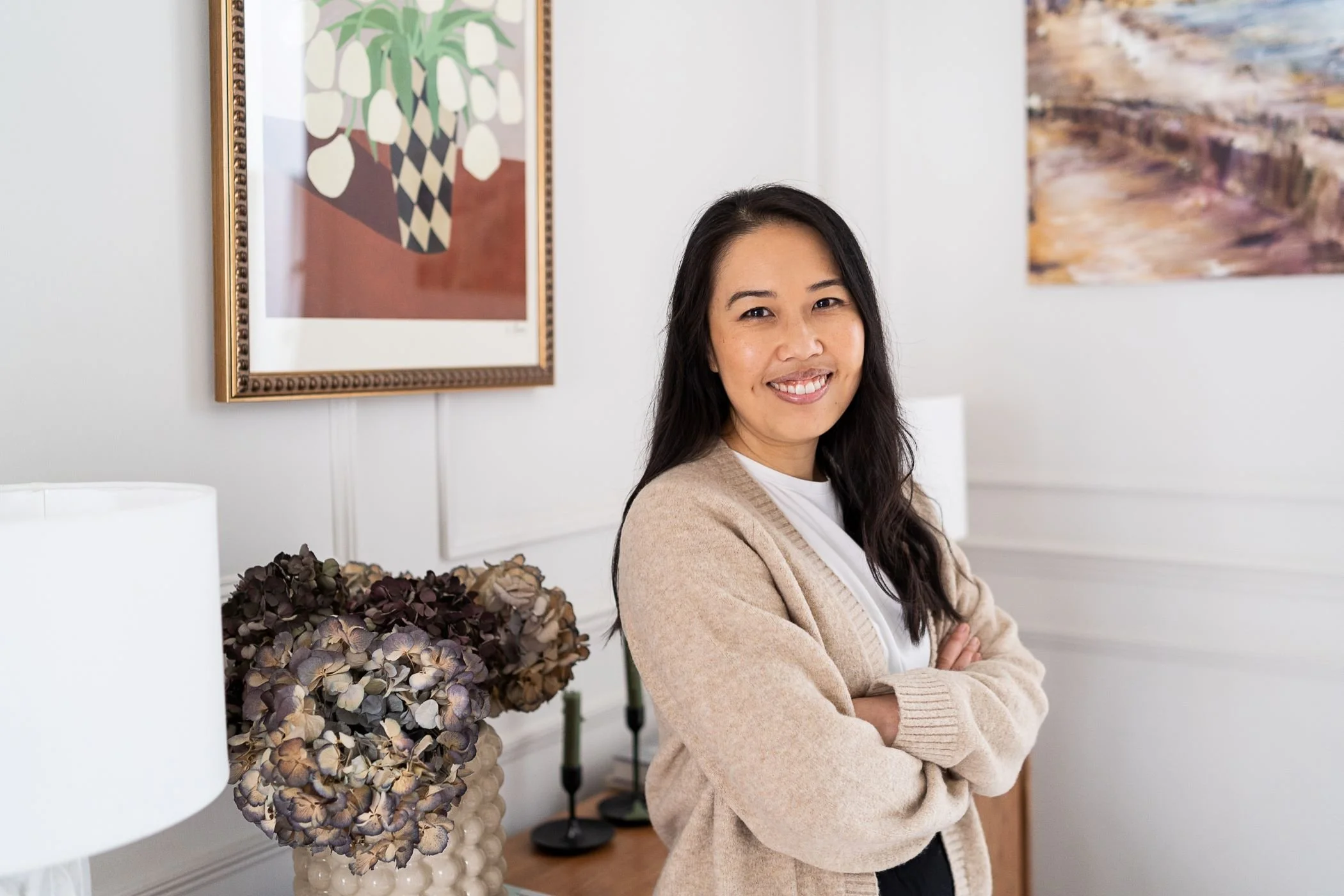 A woman with long dark hair smiling in a room with white walls, framed artwork, a vase with dried hydrangeas, and lamps.