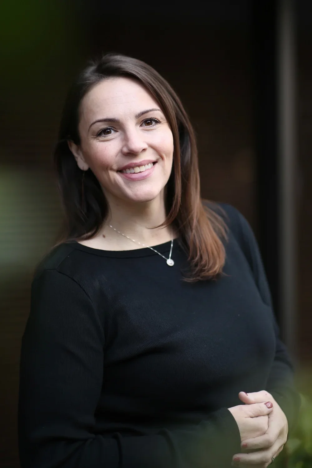 A young woman with brown hair smiling, wearing a black long-sleeve top and a silver necklace, standing outdoors at dusk.